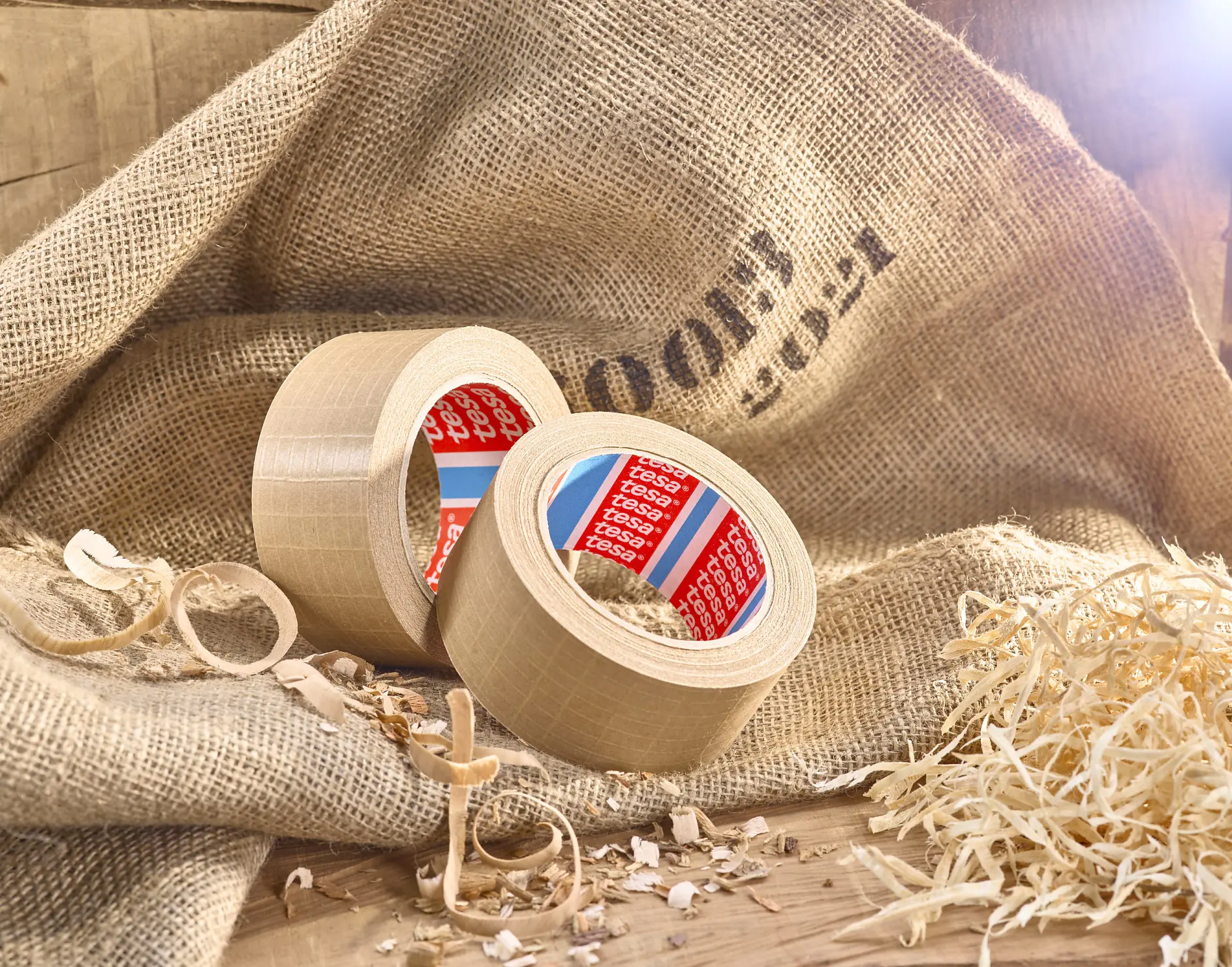 Two rolls of beige tesa tape with red inner labels sit on a burlap sack. Wood shavings are scattered around, with some curly pieces nearby. A wooden surface is in the background, and soft light illuminates the scene from the upper right.