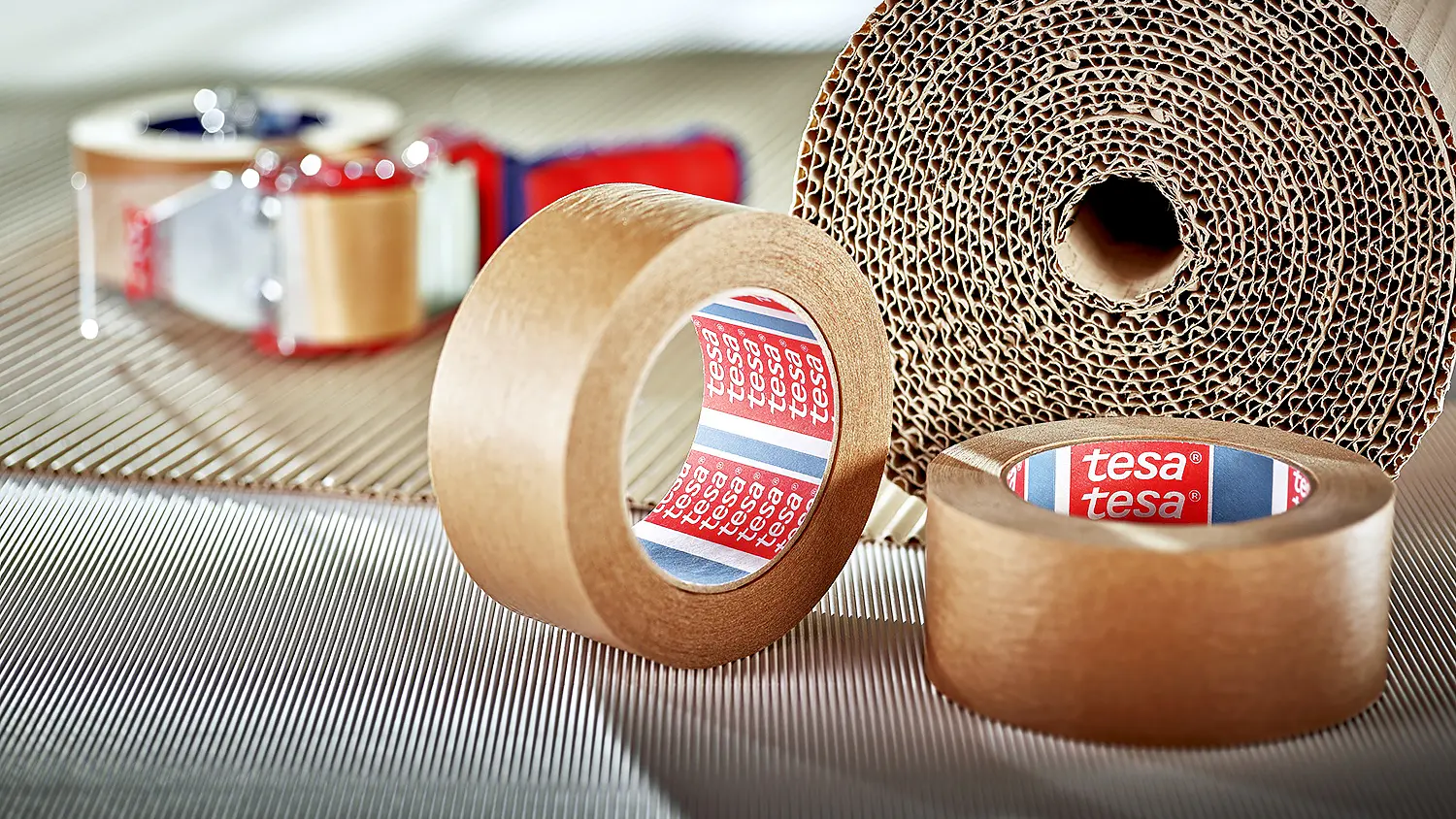 A large roll of corrugated cardboard is in the background. In the foreground, there are two smaller rolls of brown adhesive tesa tape, both labeled with the brand name. A tape dispenser with red handles is partially visible.