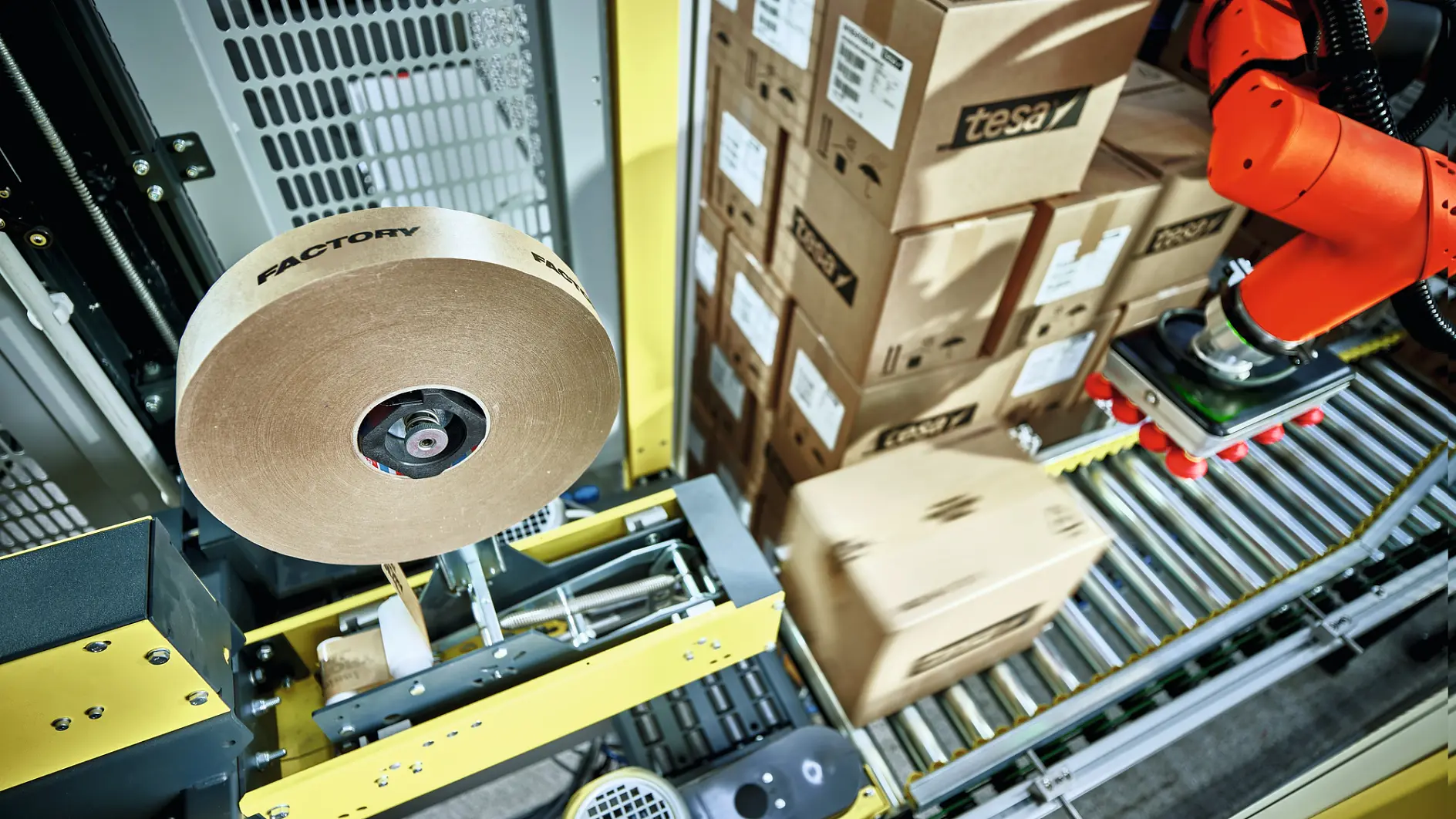 A robotic arm on a conveyor belt is stacking boxes labeled tesa. Nearby, a large roll of packing tesa tape is mounted on a machine. The setting appears to be an industrial warehouse or factory space.
