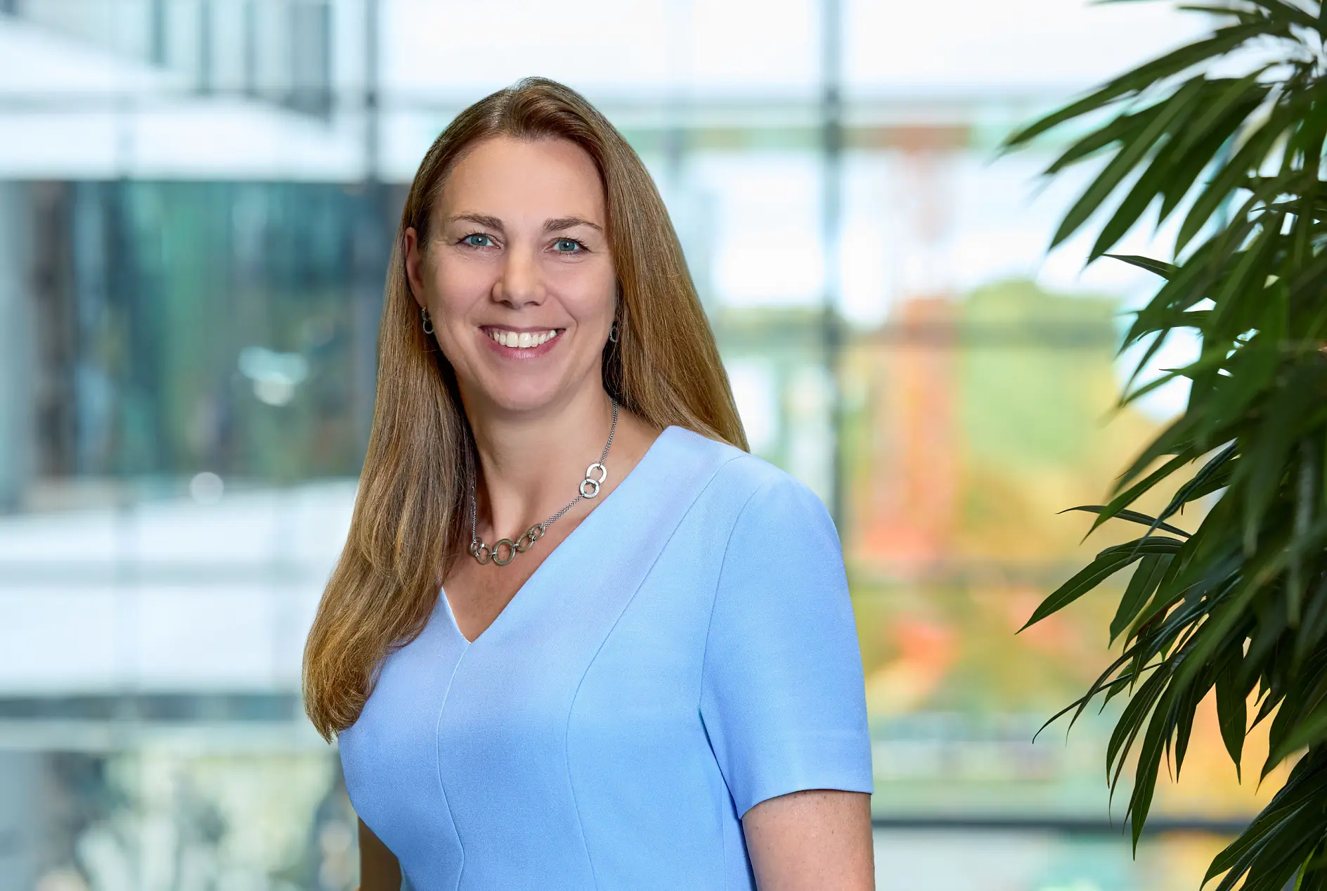 A woman with long brown hair stands in a modern hallway lined with large windows. She is wearing a black suit and is leaning on a wooden railing, which has been securely reinforced using tesa tape. The view outside shows buildings and trees. (This text has been generated by AI)