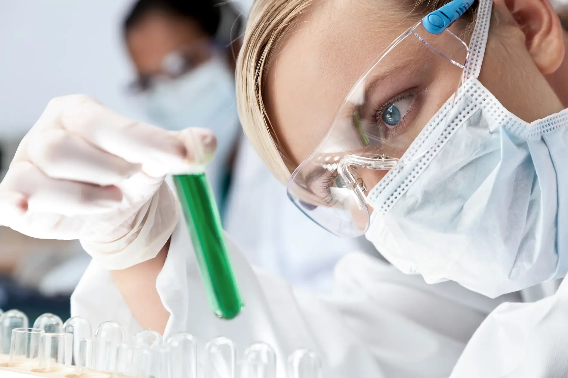 A female medical or scientific researcher or doctor looking at a green solution in a laboratory with her Asian female colleague out of focus behind her.