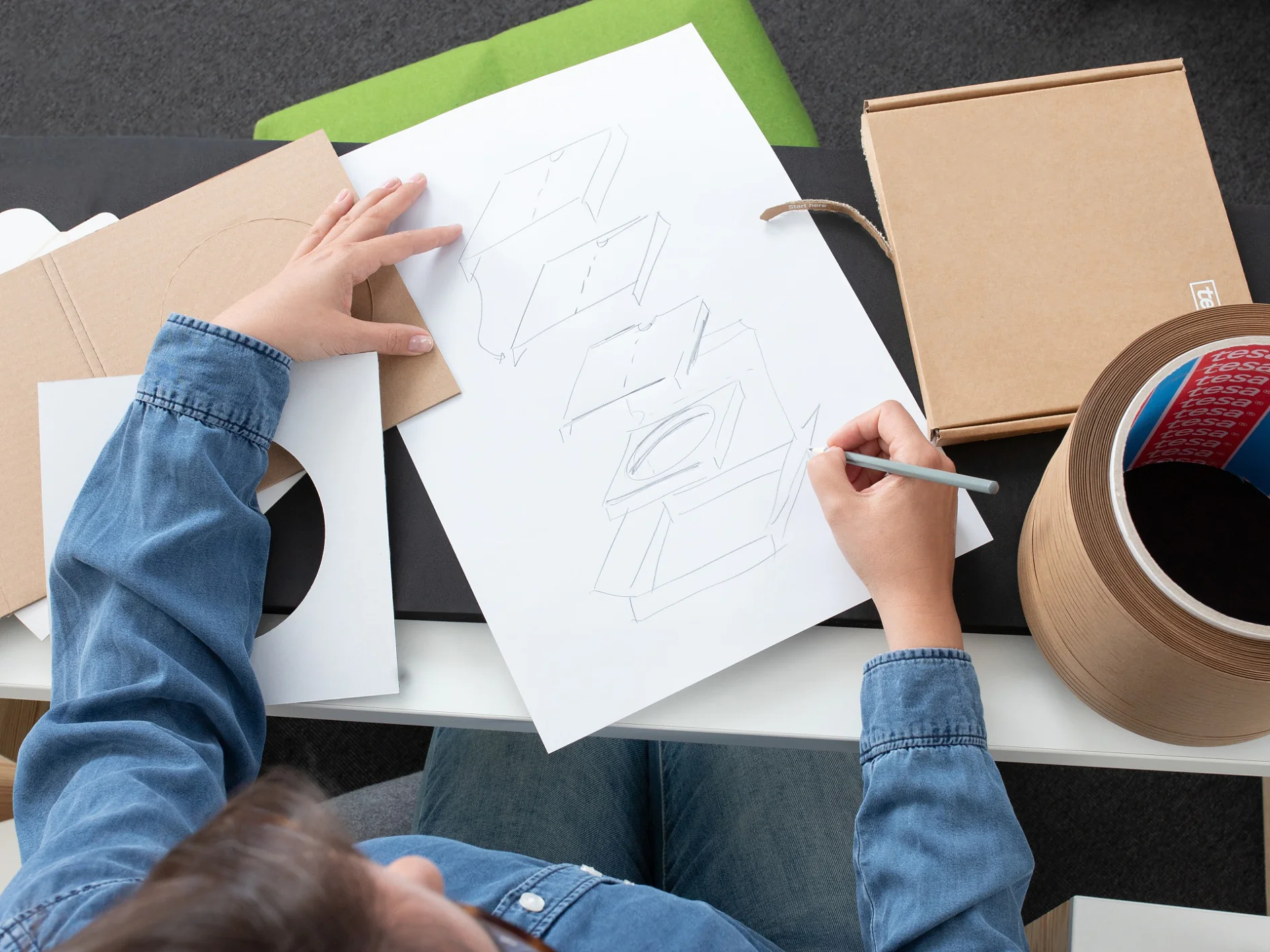 A person in a denim shirt is sketching a box design on white paper. A circular cardboard container, open cardboard box, and tesa tape are on the table. The background includes a black and white table surface with green material. (This text has been generated by AI)