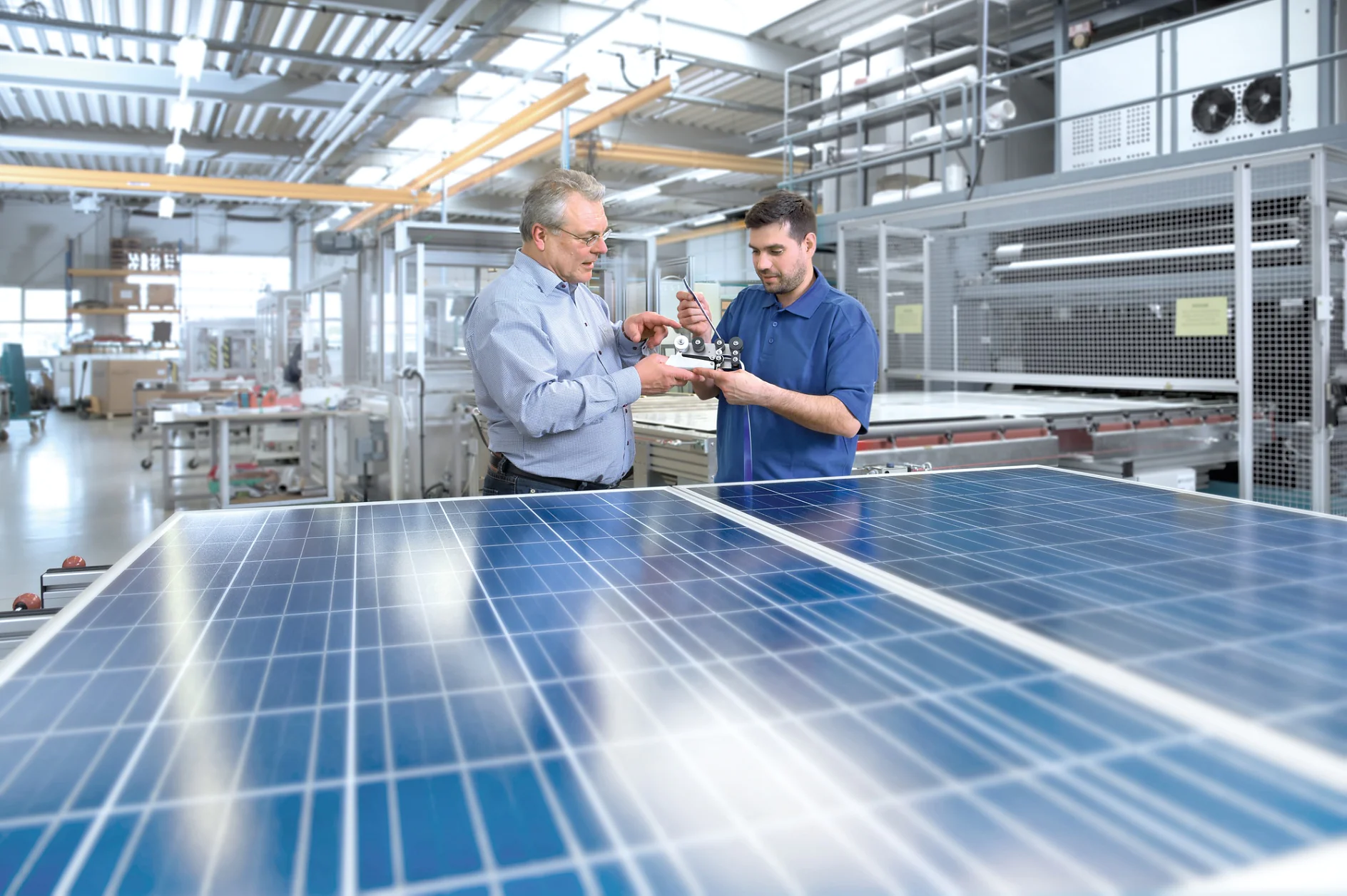 Two men are examining a small piece of machinery in a bright, industrial setting. They stand next to large solar panels. The background shows various equipment and metal structures, indicating a technological or manufacturing environment. (This text has been generated by AI)