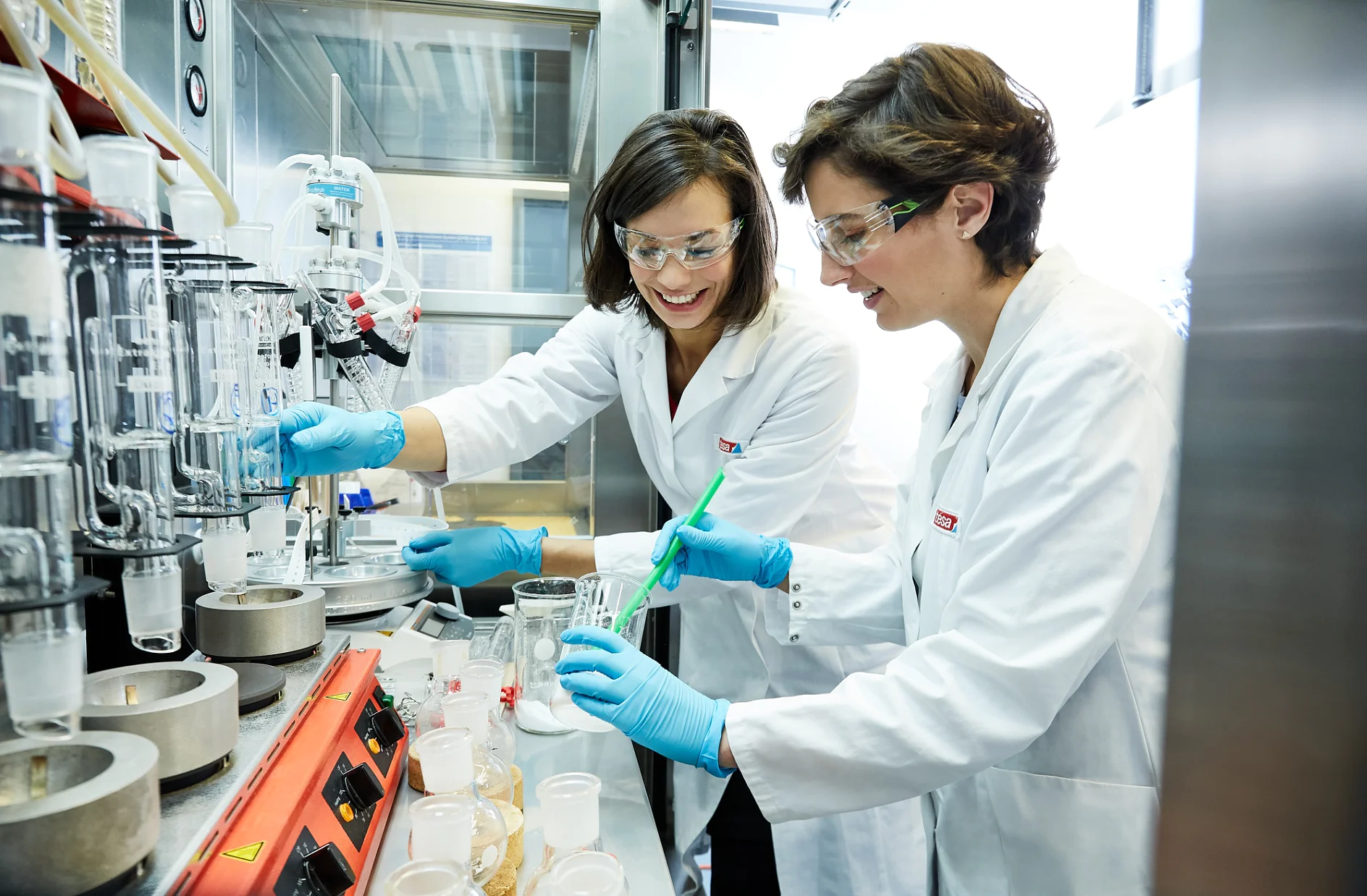 Two scientists in lab coats and safety goggles work at a laboratory bench with various equipment. They are handling chemicals, with one stirring a beaker. Both appear engaged in a research activity, utilizing tesa tape for their experimentations. (This text has been generated by AI)