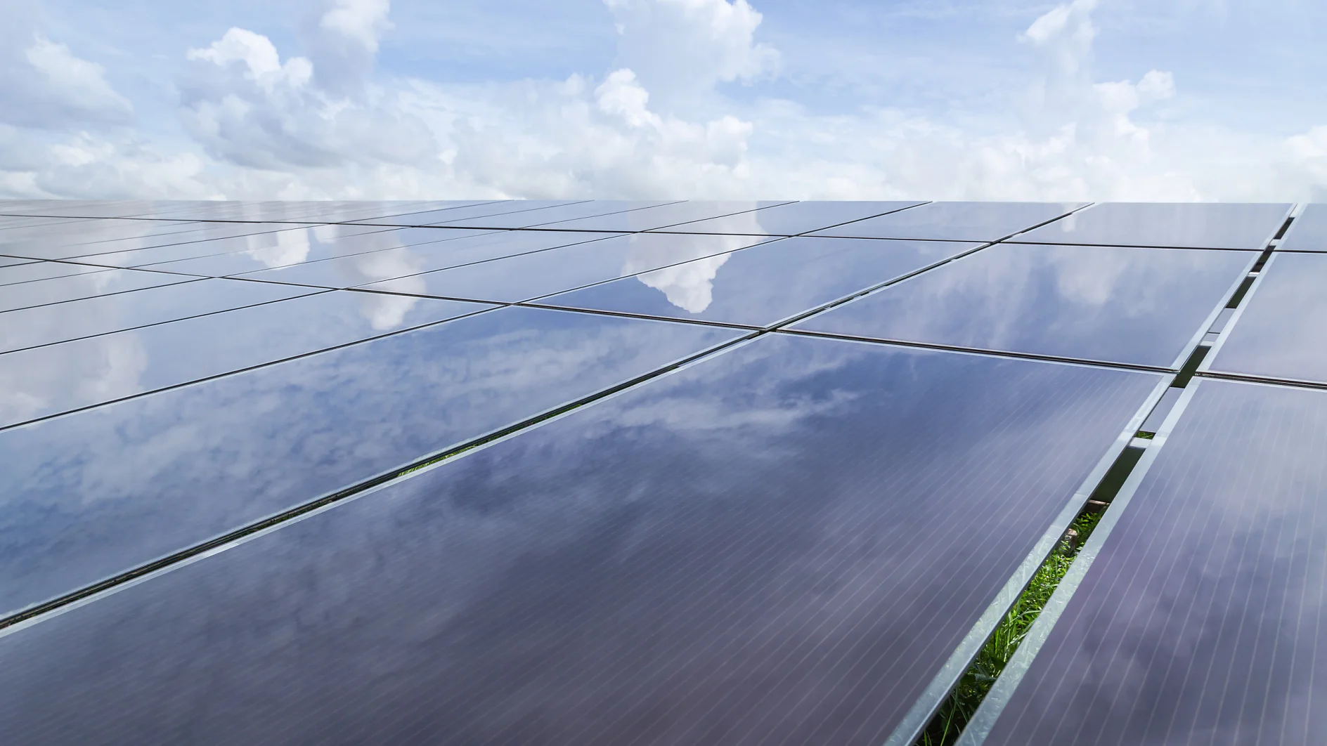 A large array of solar panels is seen reflecting a partly cloudy sky. The panels stretch across the image, capturing a mix of blue sky and white clouds. Vegetation is visible between the panels. (This text has been generated by AI)
