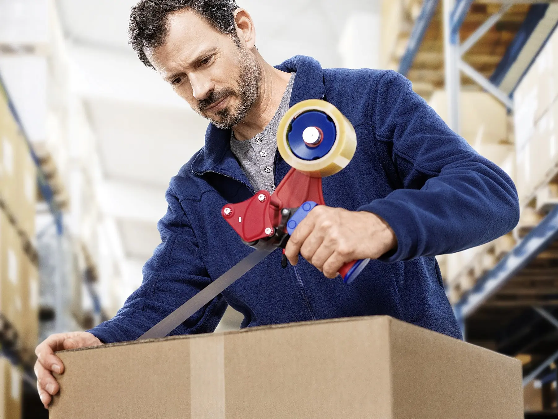 A man in a blue jacket uses a tesa tape dispenser to seal a cardboard box in a warehouse. Shelves stacked with boxes are visible in the background. (This text has been generated by AI)