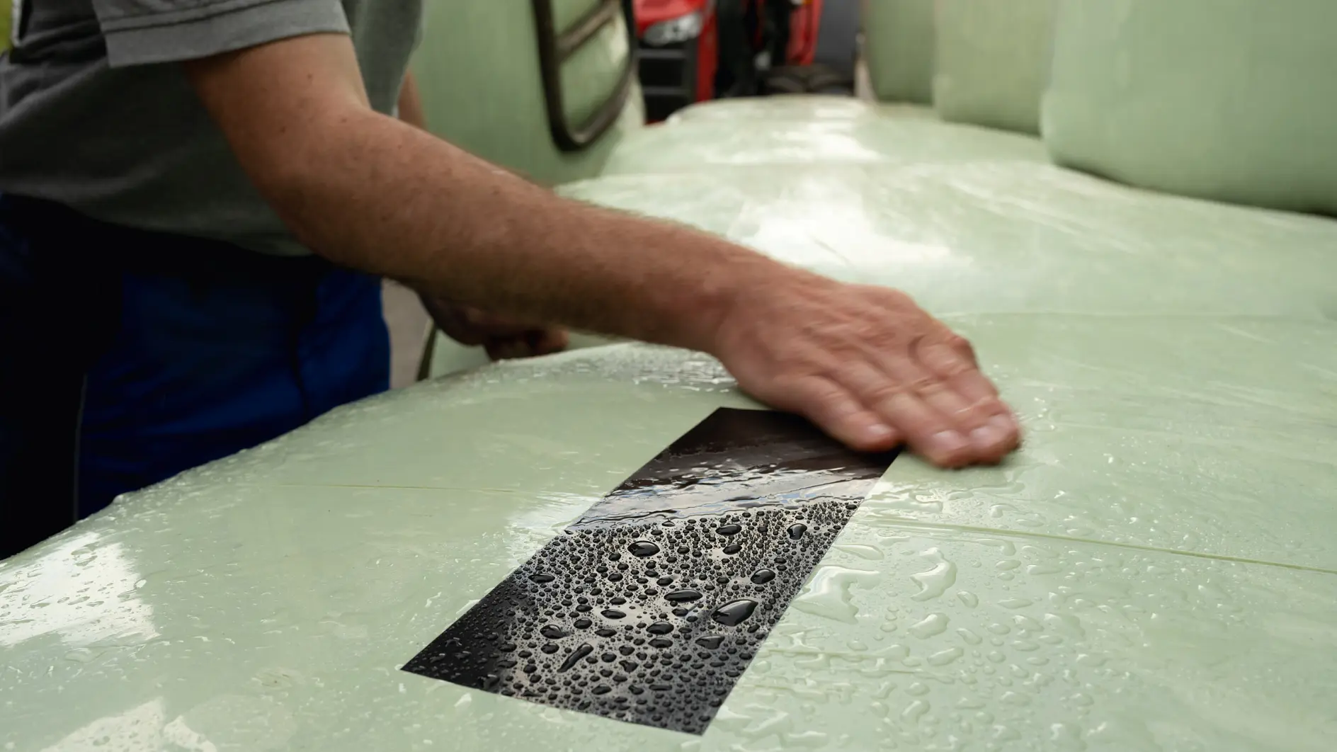 A person smooths a rectangular sticker of raindrops on a glossy, light green surface using tesa tape. They use their right hand, wearing a short-sleeve gray shirt. The sticker contrasts with the smoothness of the surrounding area.