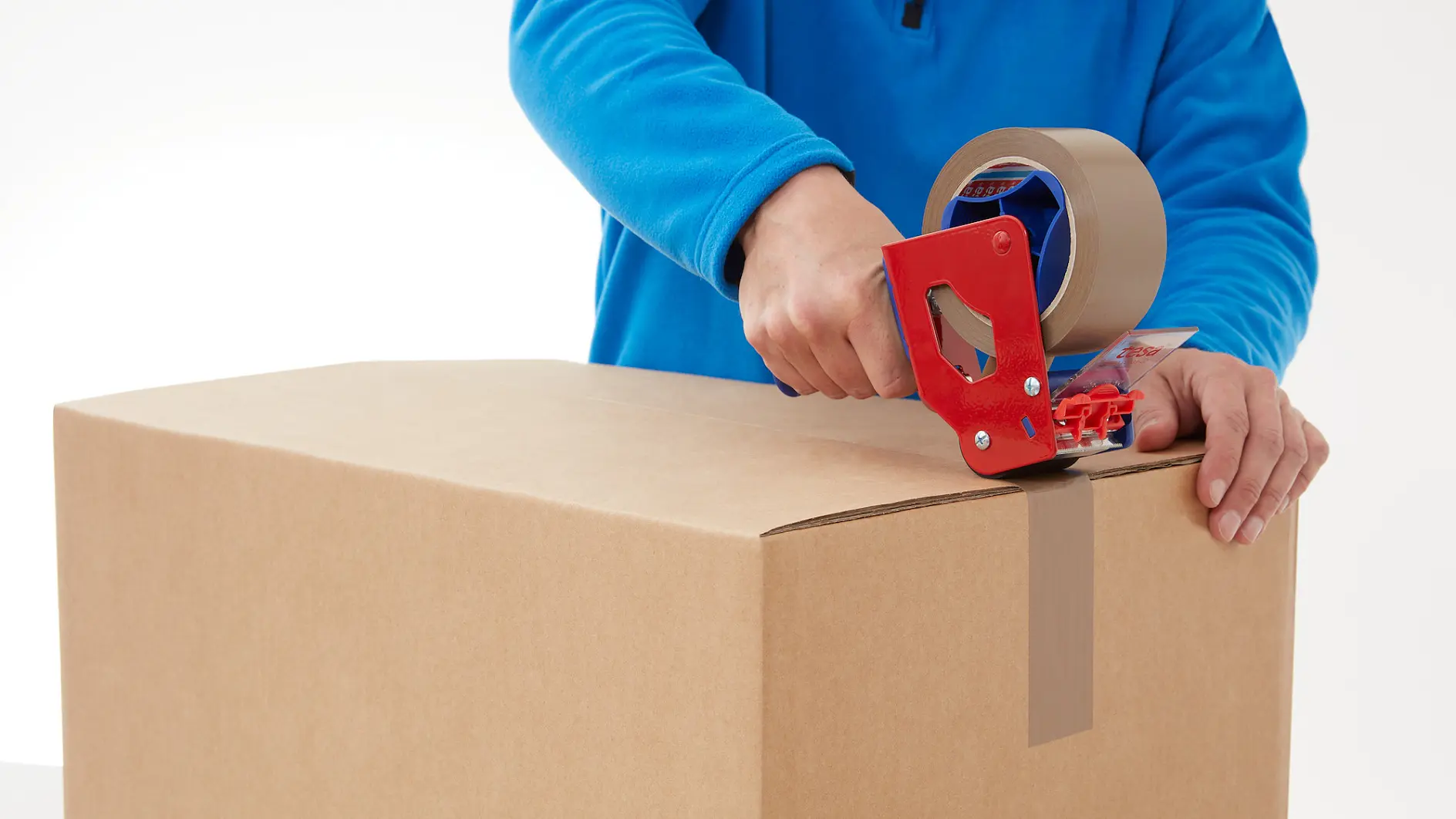 A person in a blue shirt is sealing a cardboard box using a red tape dispenser with brown tesa tape. The person is applying tesa tape along the center seam of the box. (This text has been generated by AI)