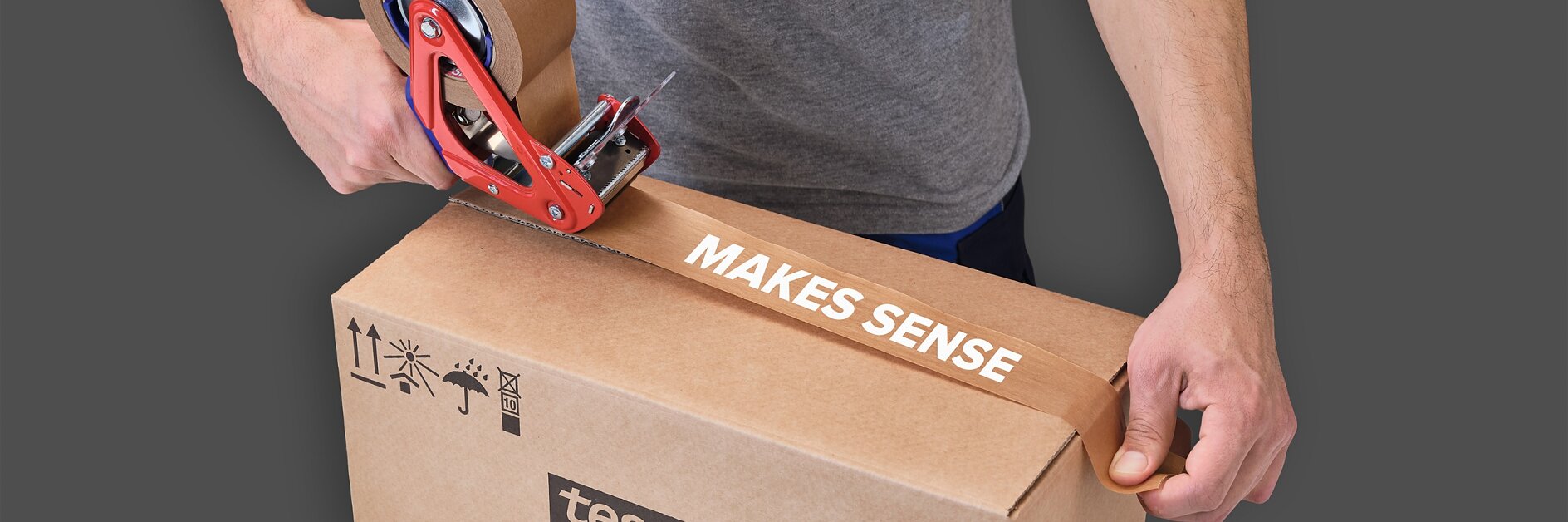 A person in a gray shirt is sealing a brown cardboard box with a red tape dispenser. The tesa tape being applied has the words MAKES SENSE printed on it. The box features the logo tesa and various handling symbols.