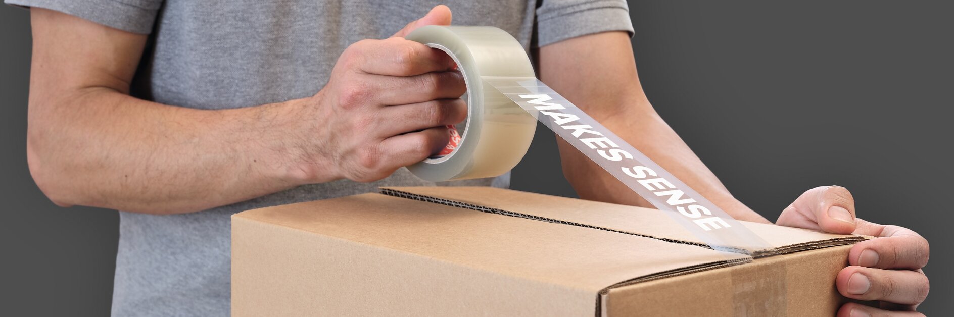 A person sealing a cardboard box using clear tesa tape with MAKES SENSE printed on it. The person is wearing a gray shirt, and the box has a tesa logo on the side.