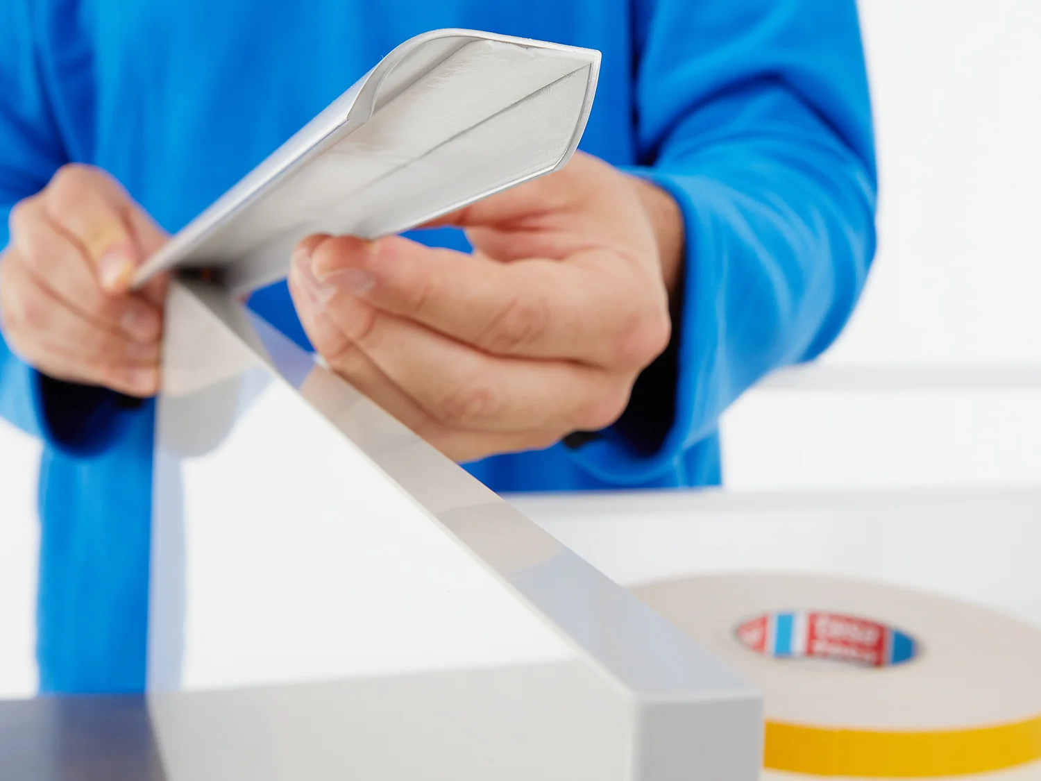 A person wearing a blue garment holds a piece of reflective tesa tape, preparing to apply it to a flat surface. A roll of similar tesa tape is visible on a white table in the foreground. (This text has been generated by AI)