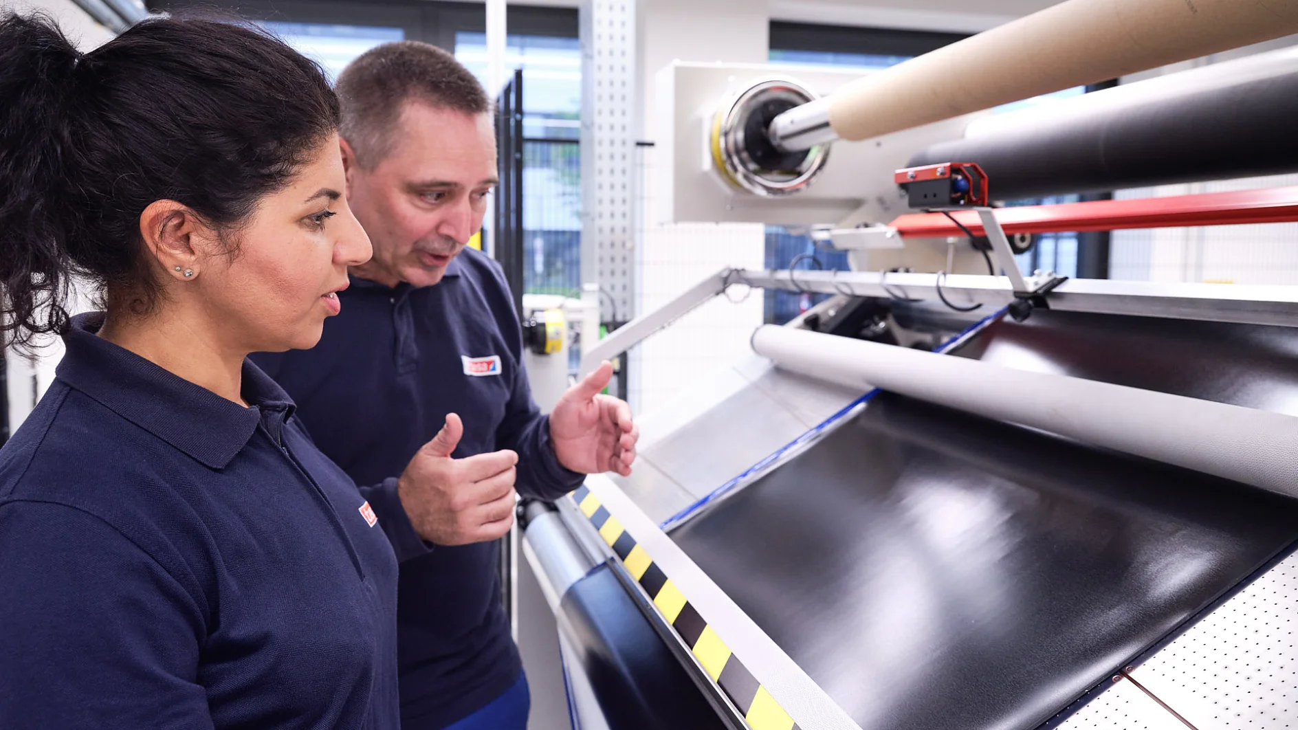 Two workers in a factory setting are observing a large sheet of black material being processed on a machine. The man explains something to the woman, demonstrating how tesa tape is used in the process. Both are wearing dark blue uniforms with red and white patches. The room is illuminated with industrial lighting. (This text has been generated by AI)