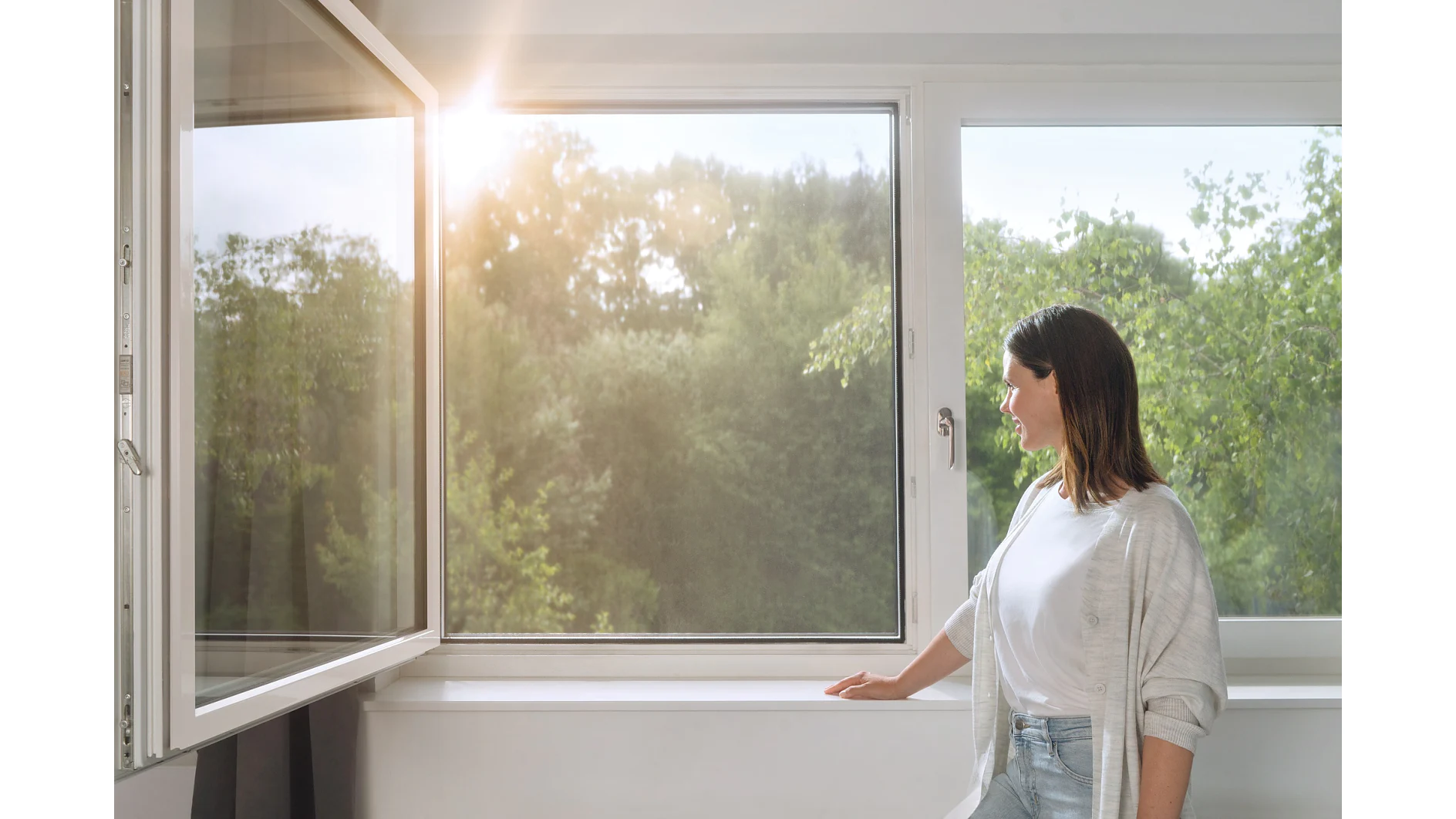 A woman stands by an open window, looking outside. Sunlight filters through, illuminating the greenery of trees in the background. She is wearing a light cardigan and jeans, with one hand resting on the windowsill secured with tesa tape. (This text has been generated by AI)