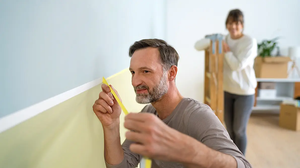 A man is carefully measuring a section of a light green wall with yellow tesa tape. Behind him, a woman is carrying a wooden step ladder. The room contains boxes and a shelf with a plant, suggesting a home improvement project.