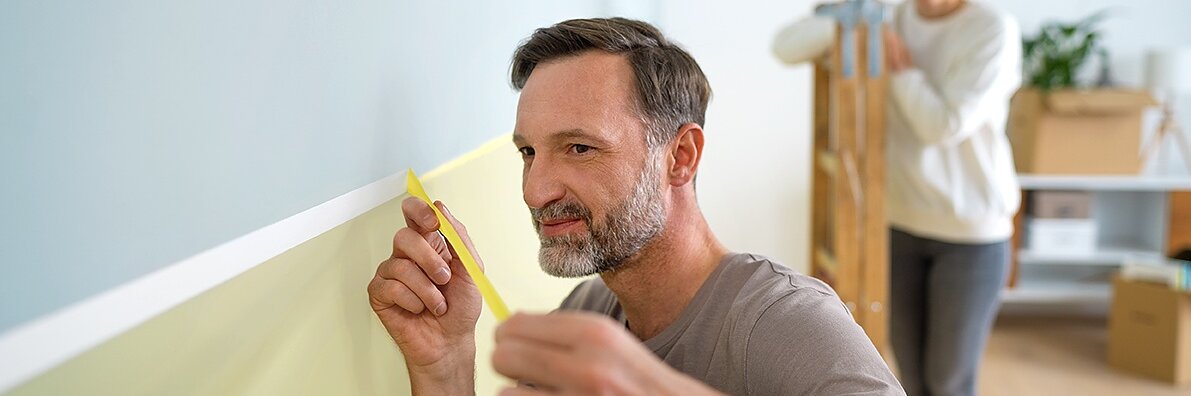 A man is carefully measuring a section of a light green wall with yellow tesa tape. Behind him, a woman is carrying a wooden step ladder. The room contains boxes and a shelf with a plant, suggesting a home improvement project.