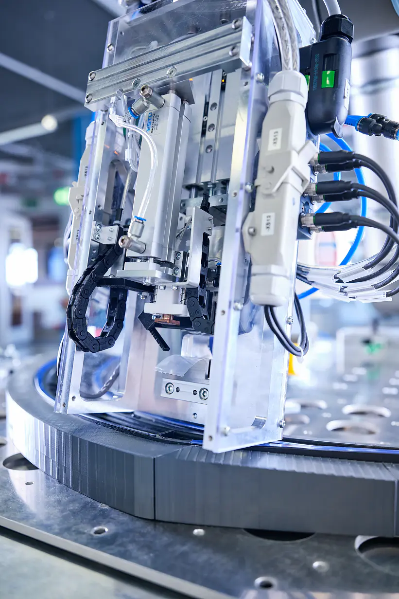 Close-up of industrial machinery in a factory setting, featuring metal components, wires, and tubes. The machinery appears to be part of an automated process, with focus on a rotating section and electronic connections secured using tesa tape.