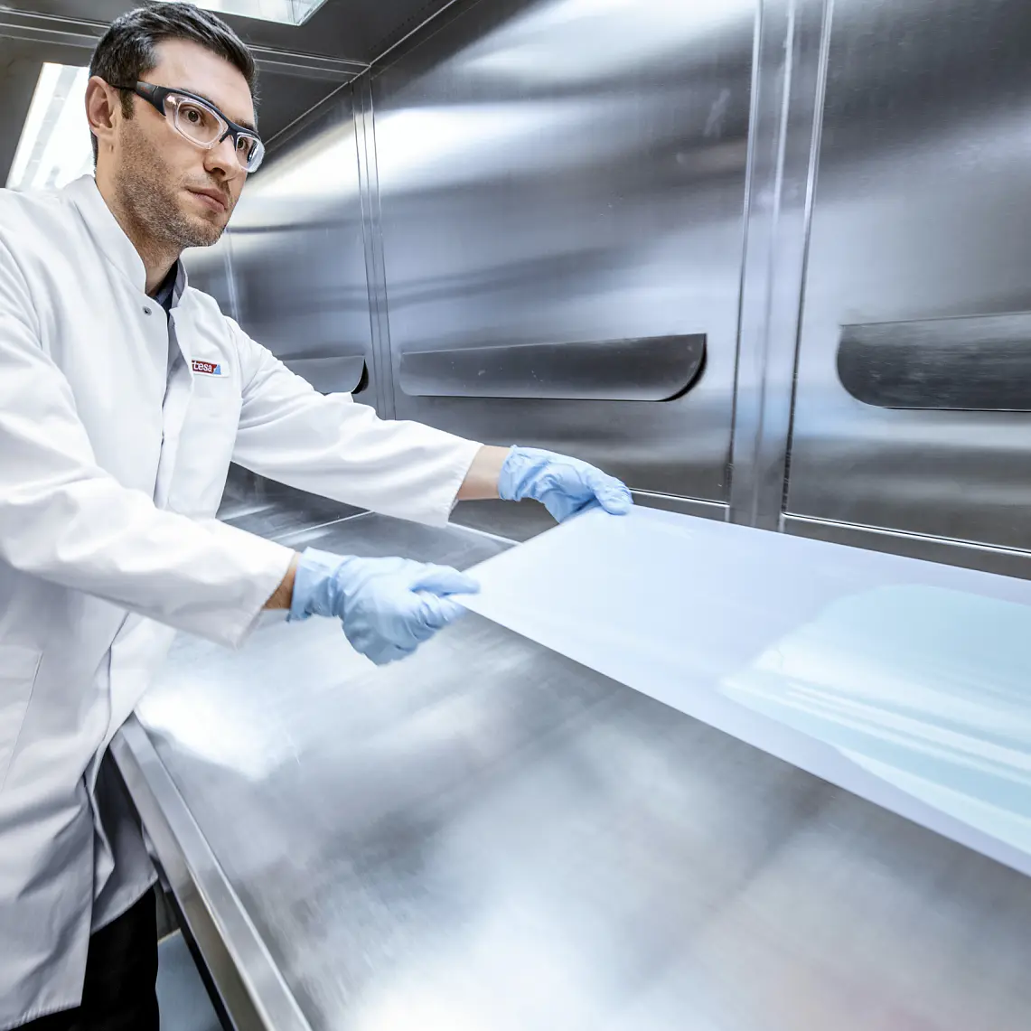 A person in a white lab coat and blue gloves works with a transparent sheet using tesa tape in a laboratory setting. The environment features metallic surfaces and bright lighting.
