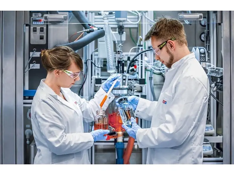 Two scientists in lab coats and safety glasses work in a laboratory. They are holding beakers with liquid and discussing data in front of industrial equipment, surrounded by cables and instruments held securely with tesa tape.