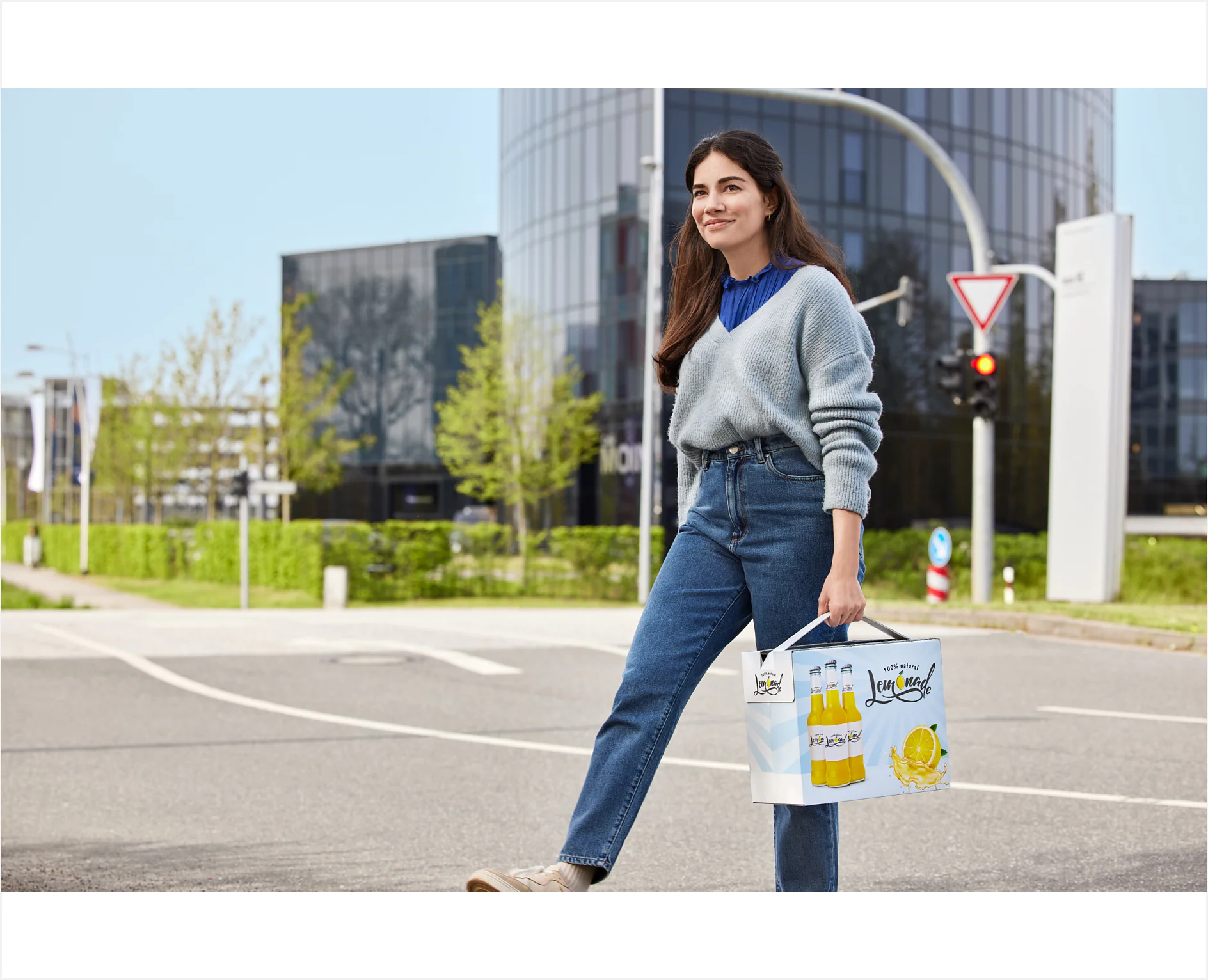 A person with long dark hair, wearing a gray sweater and blue jeans, walks across a street holding a small grocery bag sealed with tesa tape. The background features a modern glass building, greenery, and a traffic sign. (This text has been generated by AI)