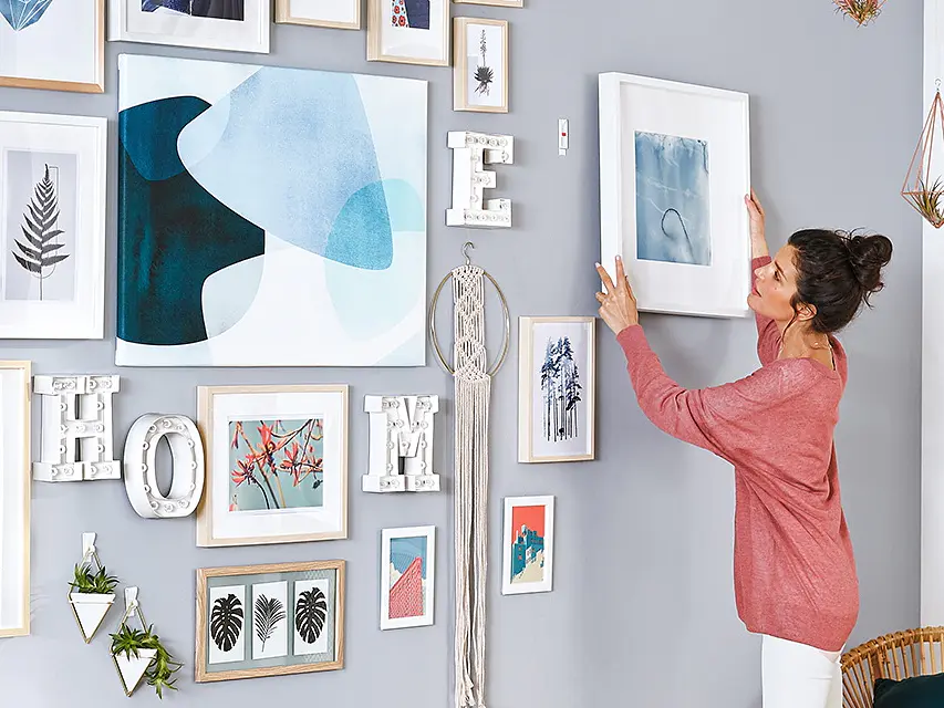 A woman in a pink top adjusts a framed picture on a gray wall filled with various artworks and decorative letters spelling HOME. The decor includes abstract pieces and nature themes in light-colored frames. Theres a wicker chair in the room. (This text has been generated by AI)