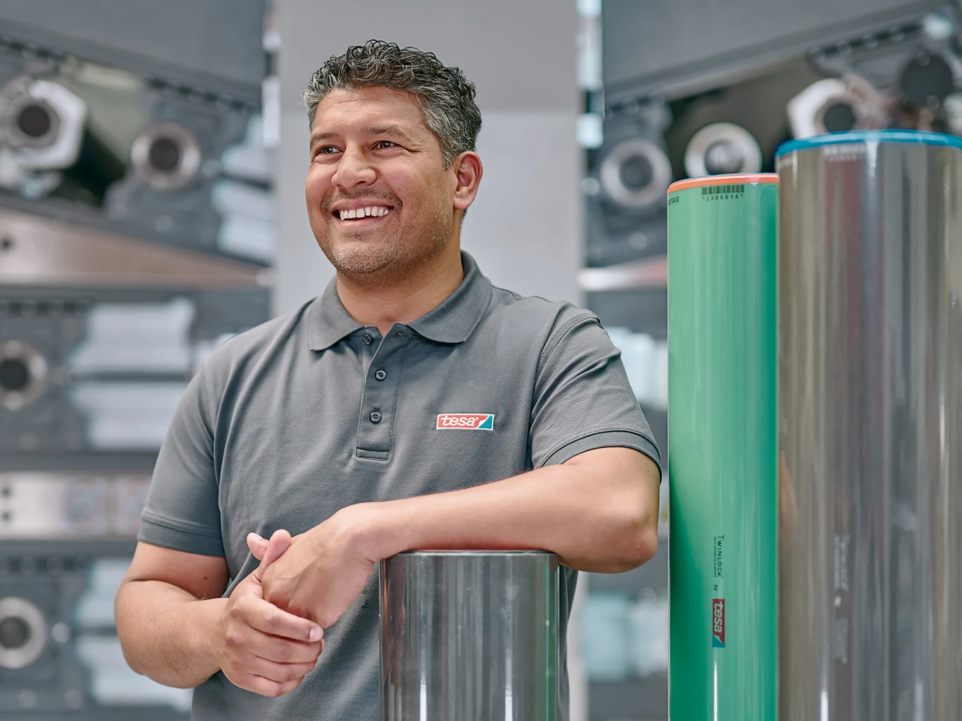 A man in a gray polo shirt stands smiling with his hands resting on a metallic cylinder in a machinery-filled environment. Two large cylindrical rolls, one metallic and one green, stand upright beside him. (This text has been generated by AI)