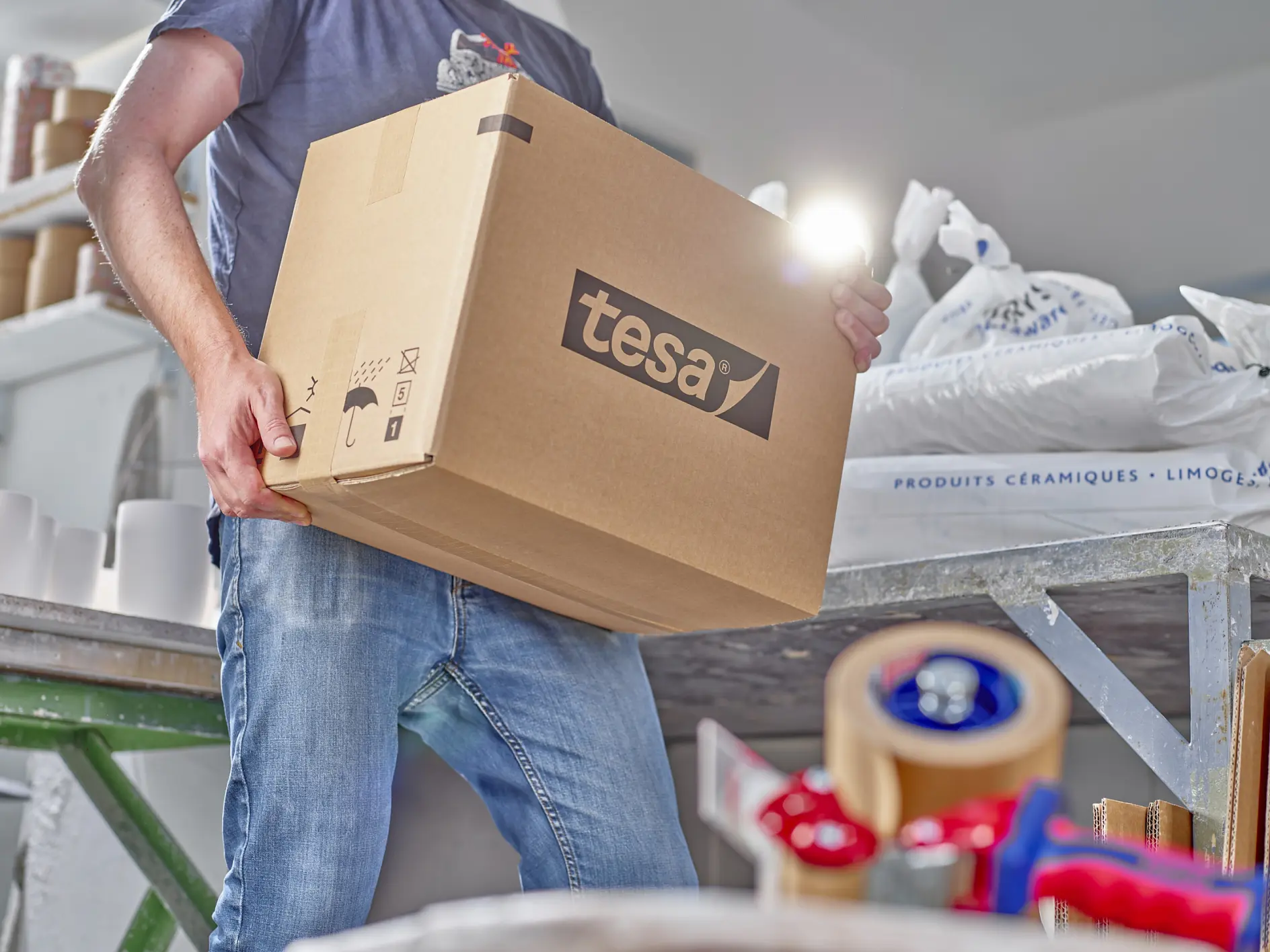 A person in a blue shirt and jeans is carrying a large cardboard box labeled tesa in a workspace. The area has packing materials, including tesa tape and rolls of paper, scattered on surfaces. (This text has been generated by AI)