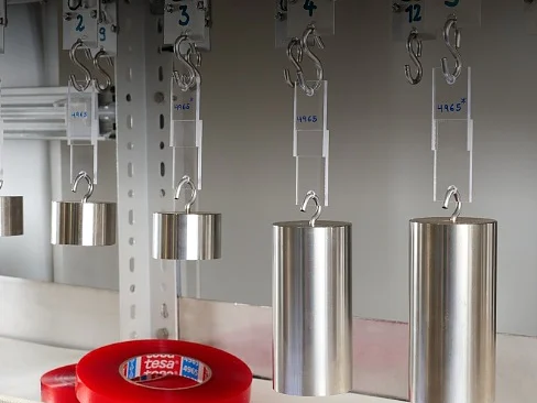 Metal weights are hanging from hooks labeled with numbers. Beneath them is a roll of red adhesive tesa tape labeled tesa. The setup appears to be part of a test or experiment environment. (This text has been generated by AI)