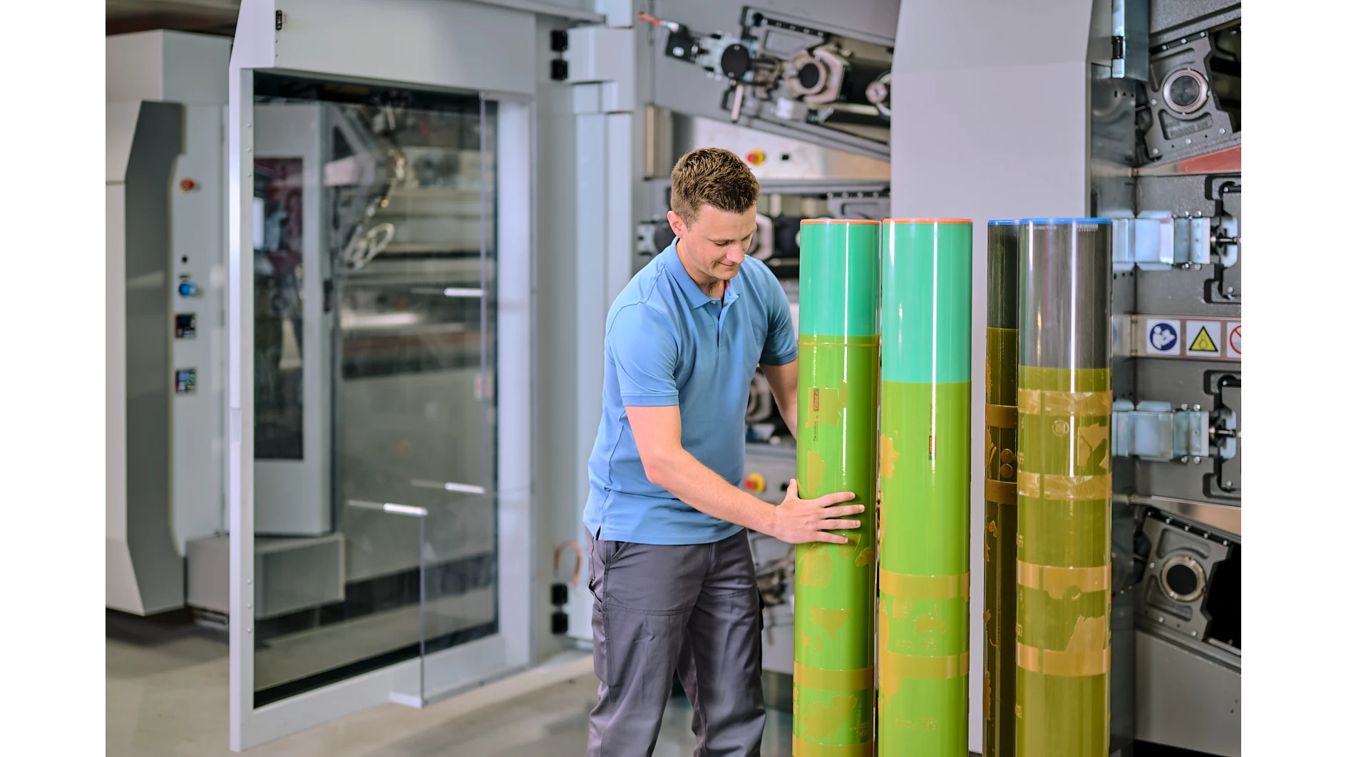A man in a blue polo shirt and gray pants is inspecting large green cylinders in an industrial setting. He stands in front of a machine with gears and panels, suggesting a manufacturing or printing environment. (This text has been generated by AI)