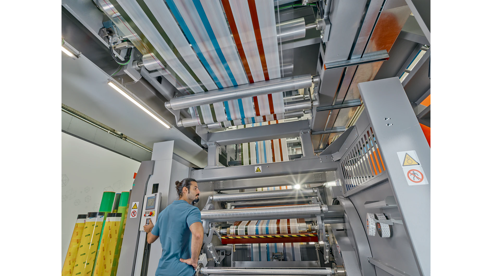 A man with a bun stands near a large industrial machine with multiple rollers, processing colorful striped material wrapped in tesa tape. The machine is metallic and has warning signs. The setting appears to be a factory or manufacturing facility. (This text has been generated by AI)