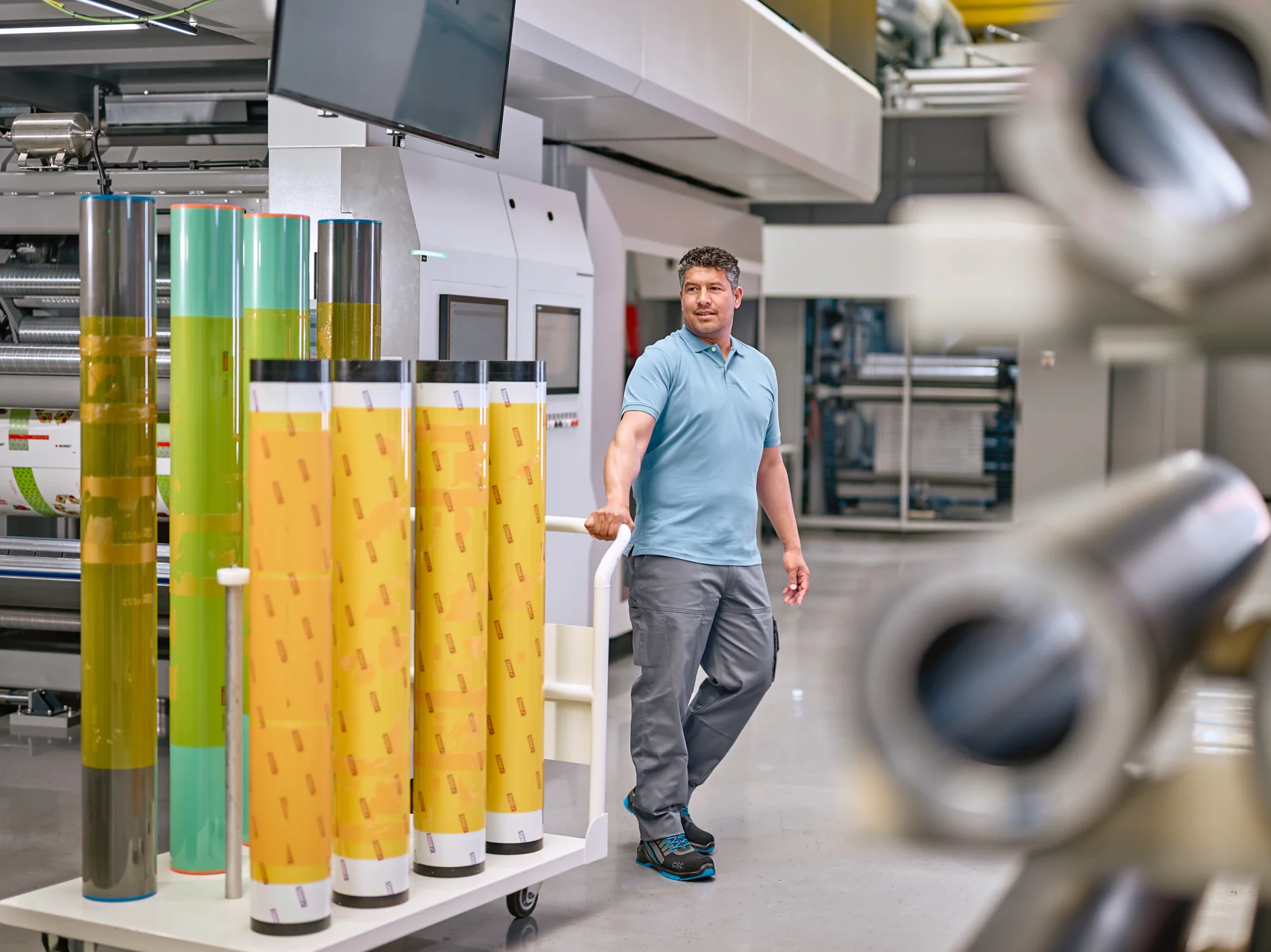 A man in a light blue polo shirt and gray pants stands in an industrial setting, surrounded by large rolls of colorful film. He rests one hand on a display of yellow and green rolls of tesa tape. Machinery and equipment are visible in the background. (This text has been generated by AI)