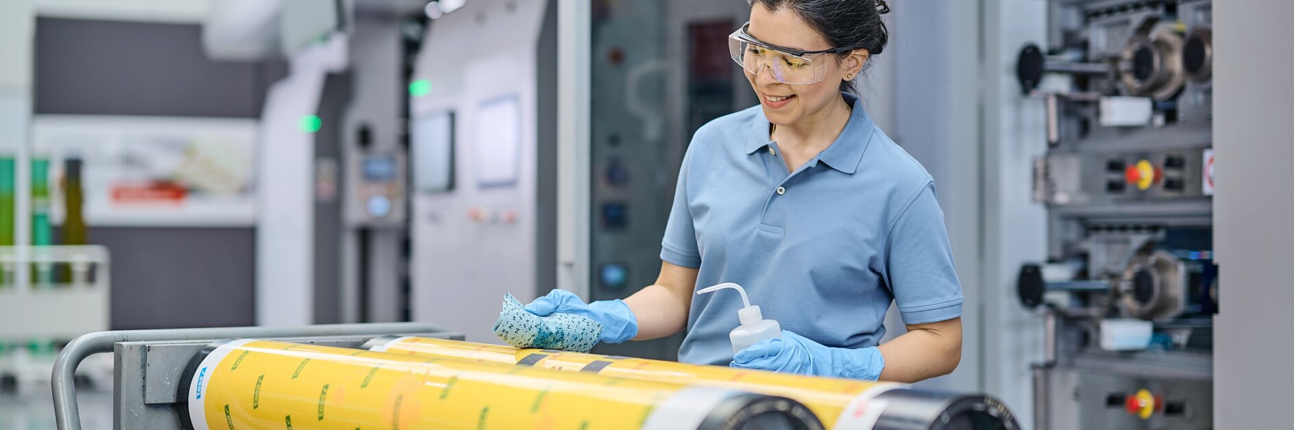 A person wearing safety glasses and gloves stands beside a printing press machine, cleaning large yellow rollers with a cloth and spray bottle. The scene appears to be in an industrial or manufacturing setting, with tesa tape nearby for additional tasks. (This text has been generated by AI)