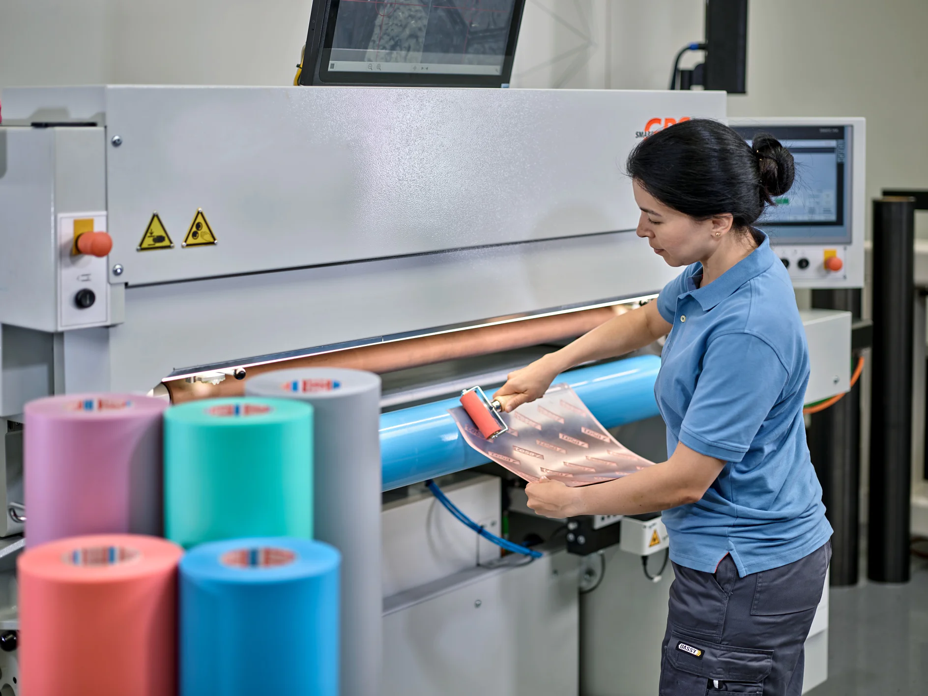 A person in a blue shirt and dark pants uses a roller on a flat surface of a large machine. The machine has screens and warning signs. Colored rolls of tesa tape are in the foreground. (This text has been generated by AI)