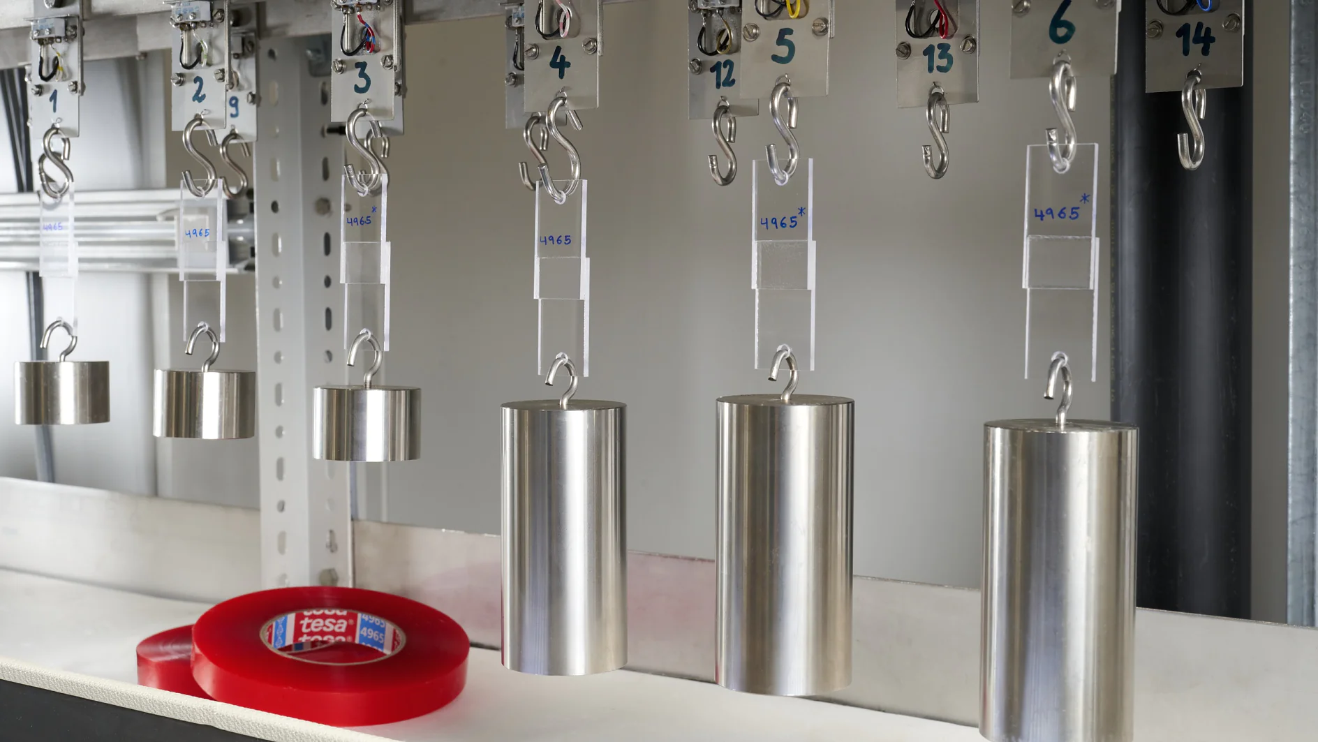 A row of metal weights hanging from hooks on a metal bar, each labeled with different numbers and text. Below them, a roll of red tesa tape is placed on a white surface. (This text has been generated by AI)