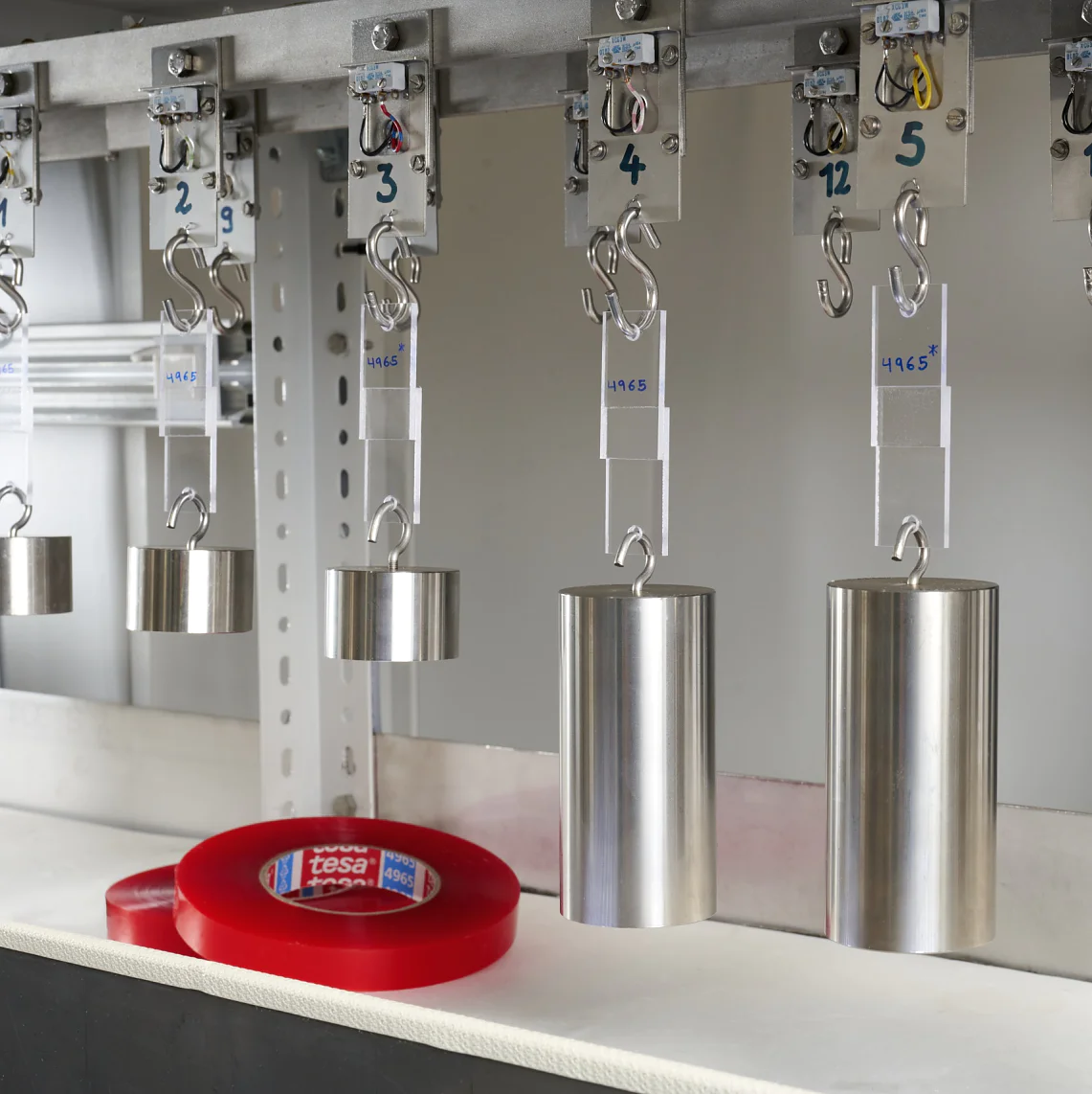 A row of metal weights hanging from hooks on a metal bar, each labeled with different numbers and text. Below them, a roll of red tesa tape is placed on a white surface. (This text has been generated by AI)