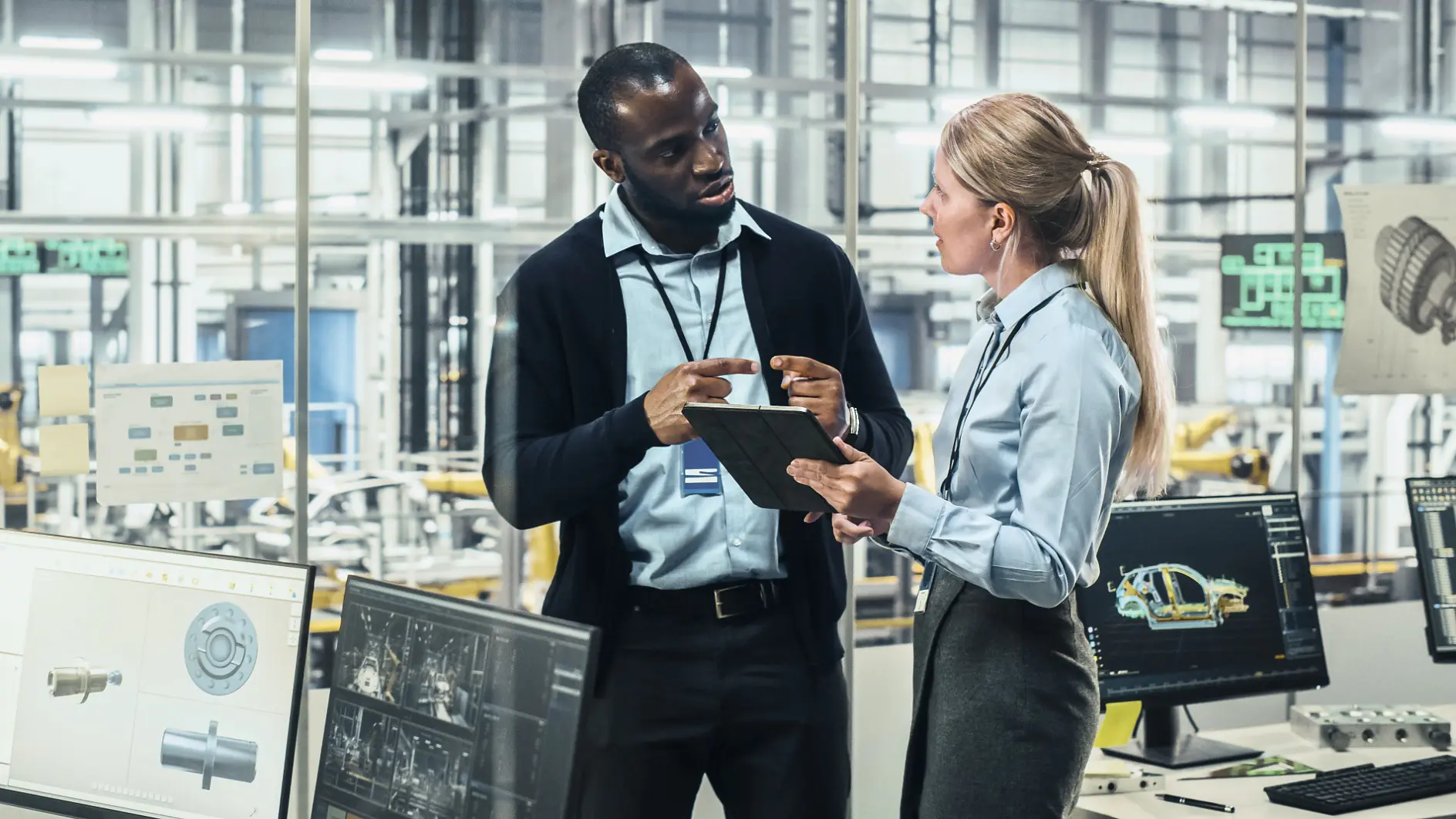 Two people stand in a factory setting, discussing work. They are surrounded by computer screens displaying technical designs and machinery. The background features industrial equipment and a large, bright workspace where tesa tape is prominently used for various applications. (This text has been generated by AI)