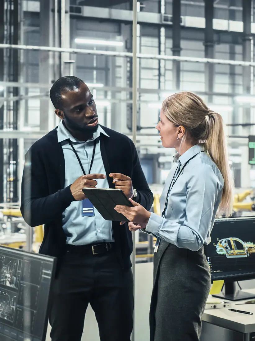 Two people stand in a factory setting, discussing work. They are surrounded by computer screens displaying technical designs and machinery. The background features industrial equipment and a large, bright workspace where tesa tape is prominently used for various applications. (This text has been generated by AI)