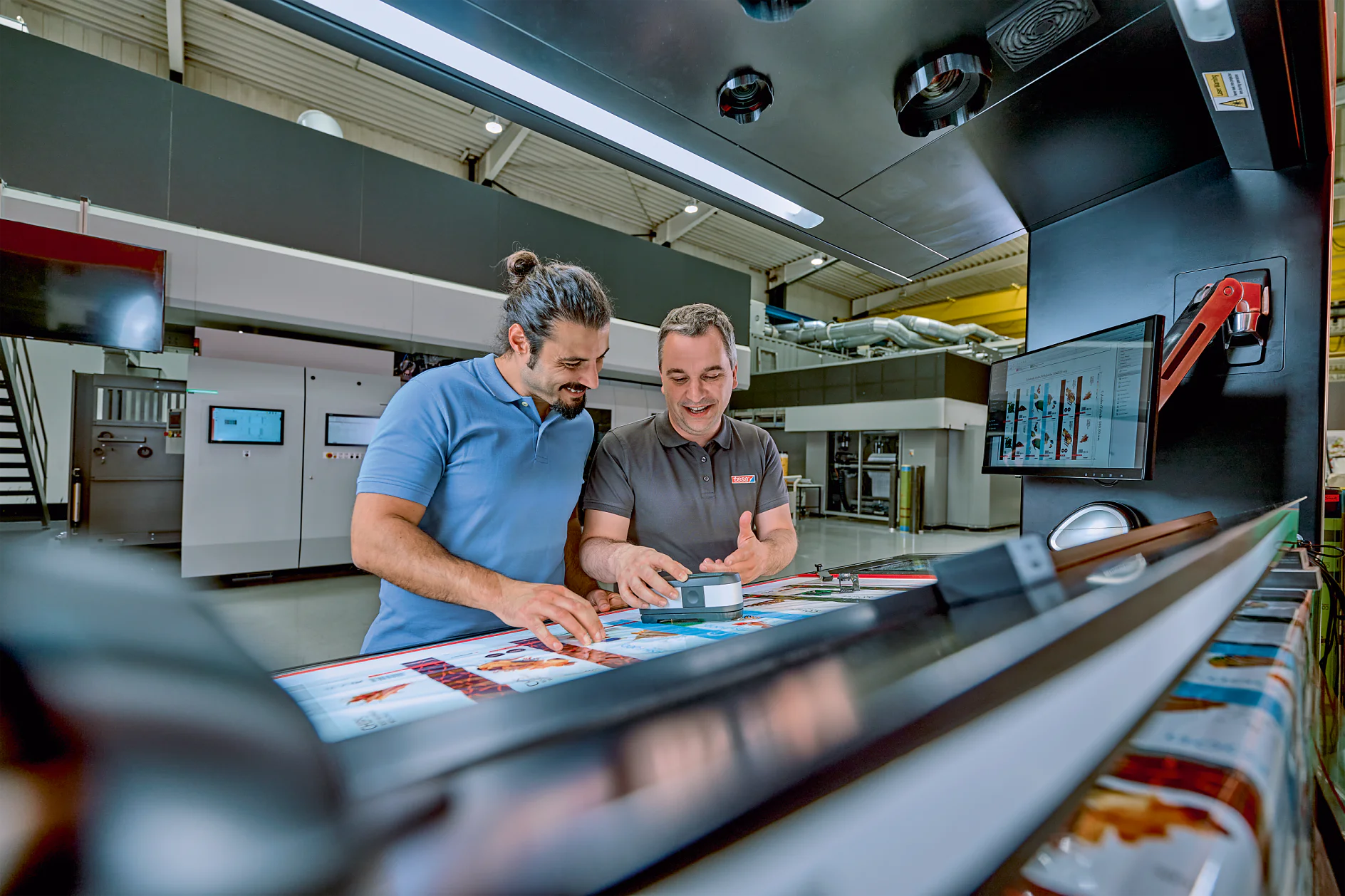 Two men are standing in a modern printing facility, examining colorful printed materials on a large machine. One man is holding a small box of tesa tape and smiling. There are screens and equipment in the background. (This text has been generated by AI)