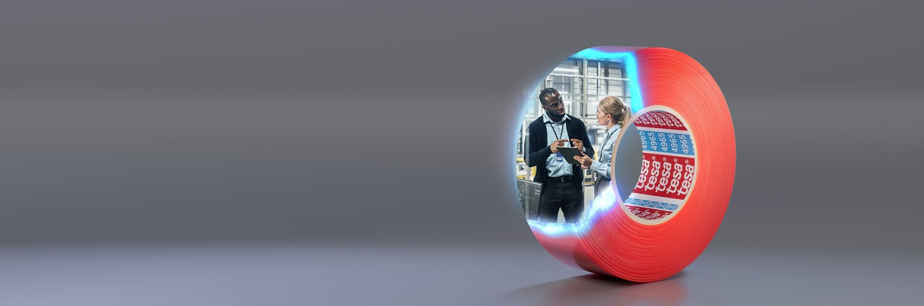 A large roll of red tesa tape stands upright on a gray surface. The front of the roll shows an image of two people discussing something, and behind them is industrial equipment. The inner side of the roll has rows of blue and red cards.