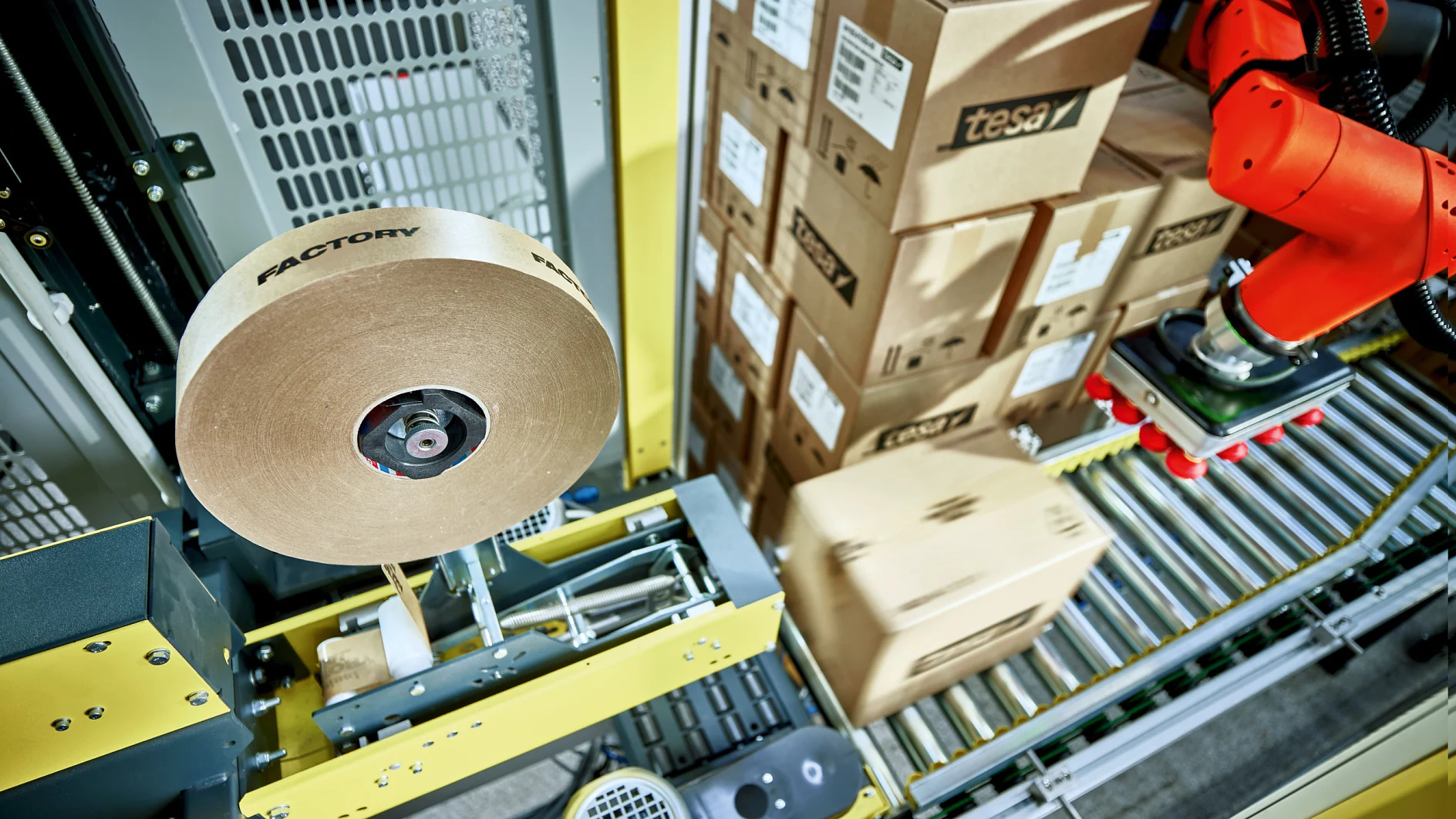A robotic arm is operating in a factory setting, handling cardboard boxes on a conveyor system. A roll of tesa tape labeled factory is part of the machinery. Stacked boxes in the background are marked with tesa. (This text has been generated by AI)