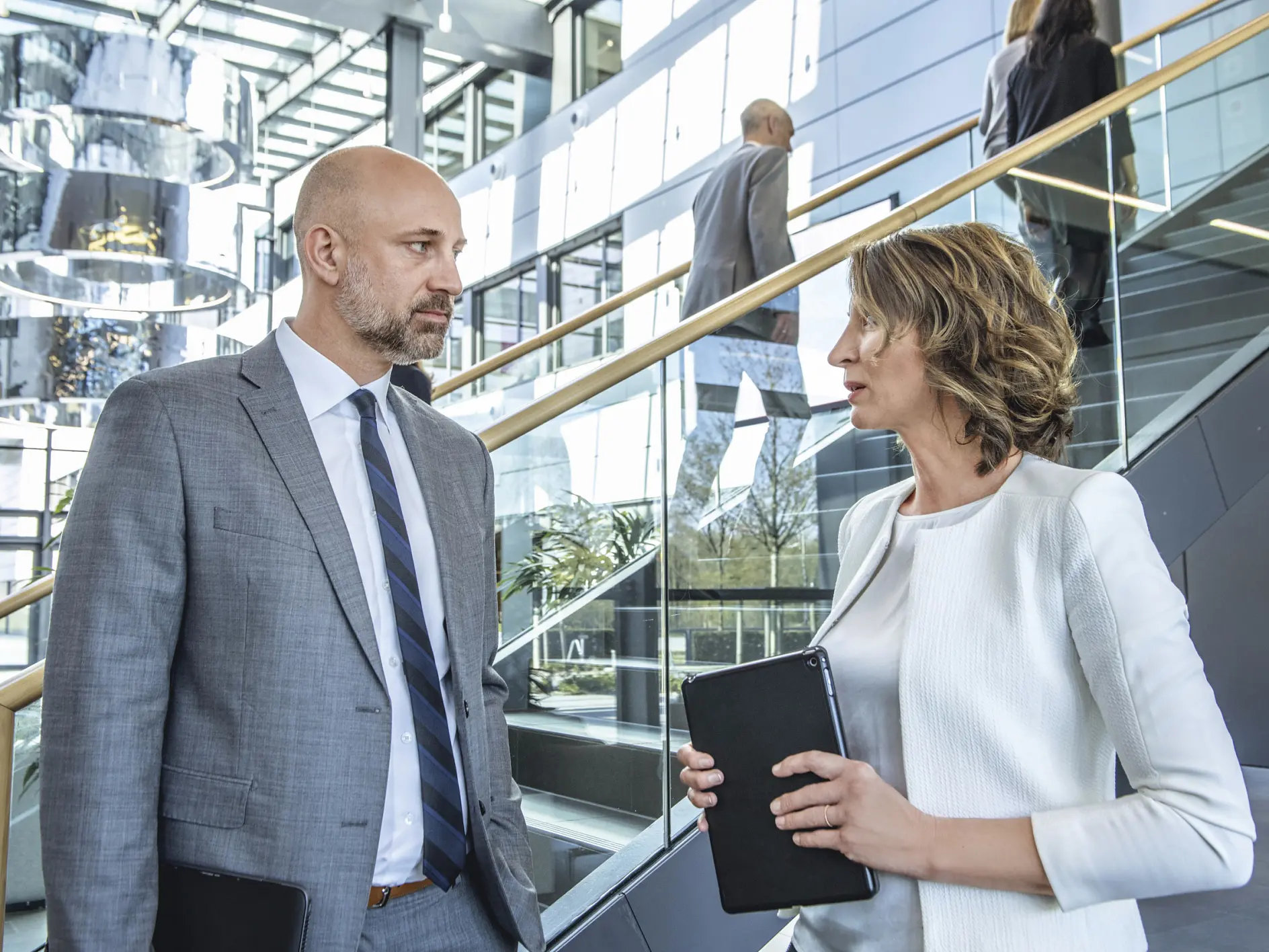 A man in a gray suit and a woman in a white jacket are conversing while holding tablets. They are standing near a modern staircase with glass railings in an office environment. Two other people, who might also be chatting about their favorite brand of tesa tape, are visible in the background.