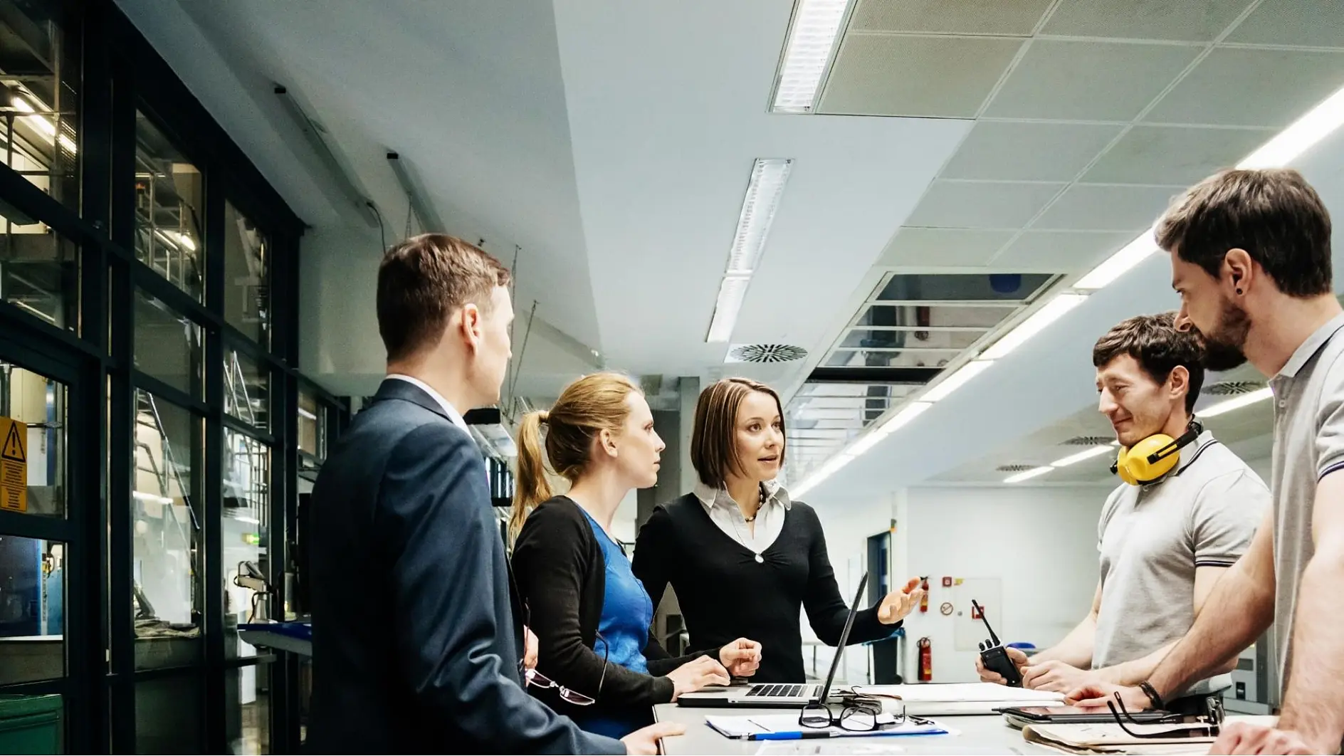 Five people are having a discussion around a desk in a modern office. One woman is gesturing while speaking, with tesa tape and a laptop in front of her. Two people are wearing casual clothing, and one is wearing yellow headphones around his neck.