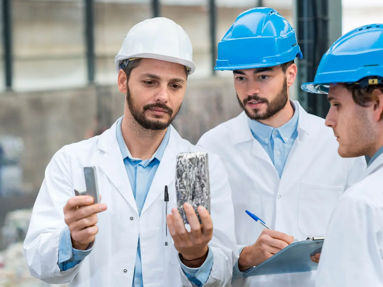 Three men wearing white lab coats and hard hats examine a rectangular object wrapped in tesa tape. Two men hold notebooks, and one is taking notes. They are standing in an industrial or recycling facility. (This text has been generated by AI)