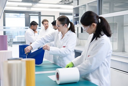 Laboratuvar önlükleri ve güvenlik gözlükleri takan birkaç kişi, arka planda raflar ve laboratuvar ekipmanlarının bulunduğu bir laboratuvar çalışma alanında büyük tesa bant ruloları ve kesici aletlerle çalışıyor.