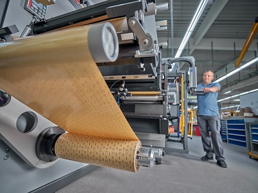 Bir adam, parlak bir fabrika ortamında, içine kahverengi tesa bant rulosu beslenen büyük bir endüstriyel makinenin yanında duruyor.