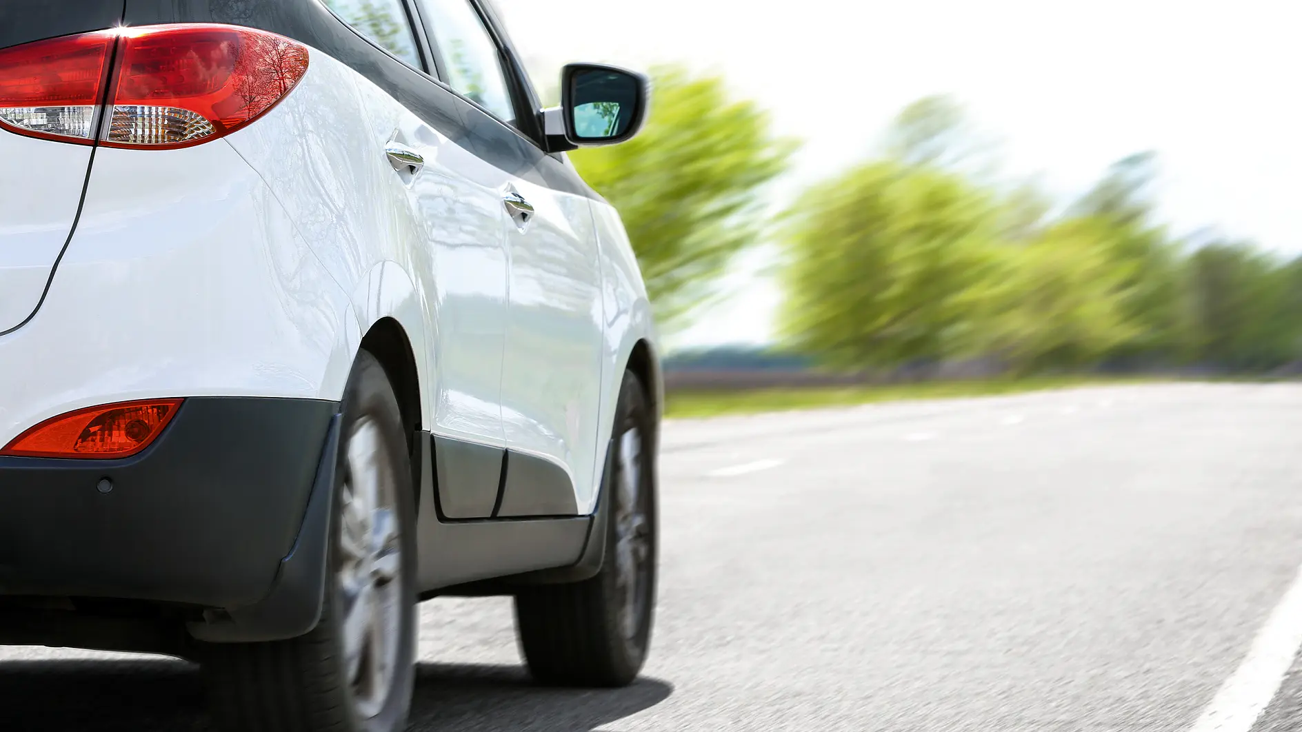 white compact car driving on a paved road with blurred green trees in the background on a clear day (This text has been generated by AI)