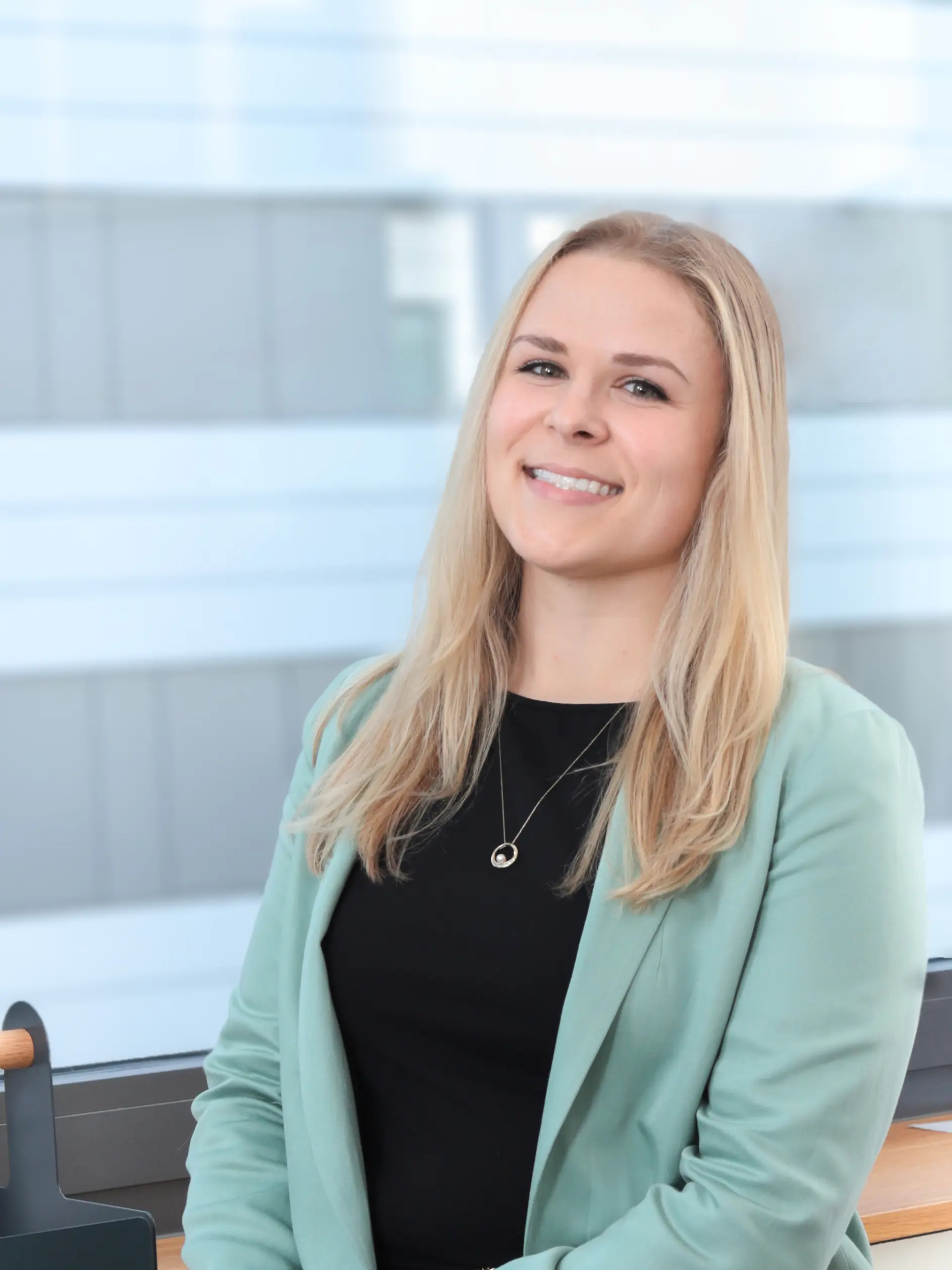 Uzun sarı saçlı, nane yeşili blazer ceket ve siyah bluz giymiş bir kadın içeride, pencere kenarında oturuyor ve kameraya hafifçe gülümsüyor. (This text has been generated by AI)