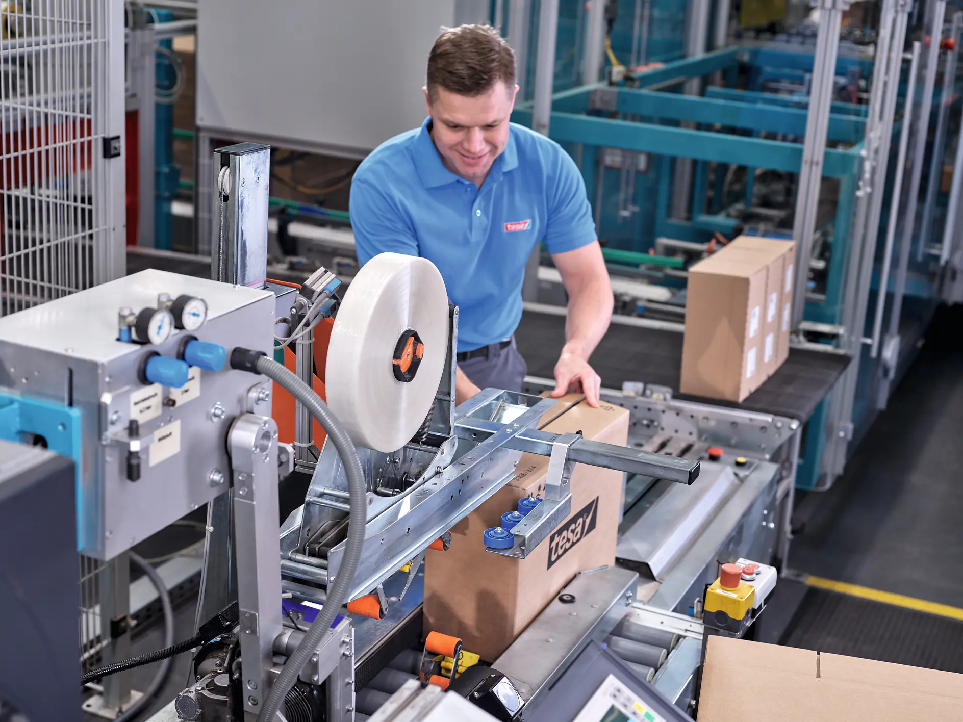 mavi tesa tişört giyen işçi, endüstriyel depo ortamında karton kutularla paketleme makinesi çalıştırıyor (This text has been generated by AI)