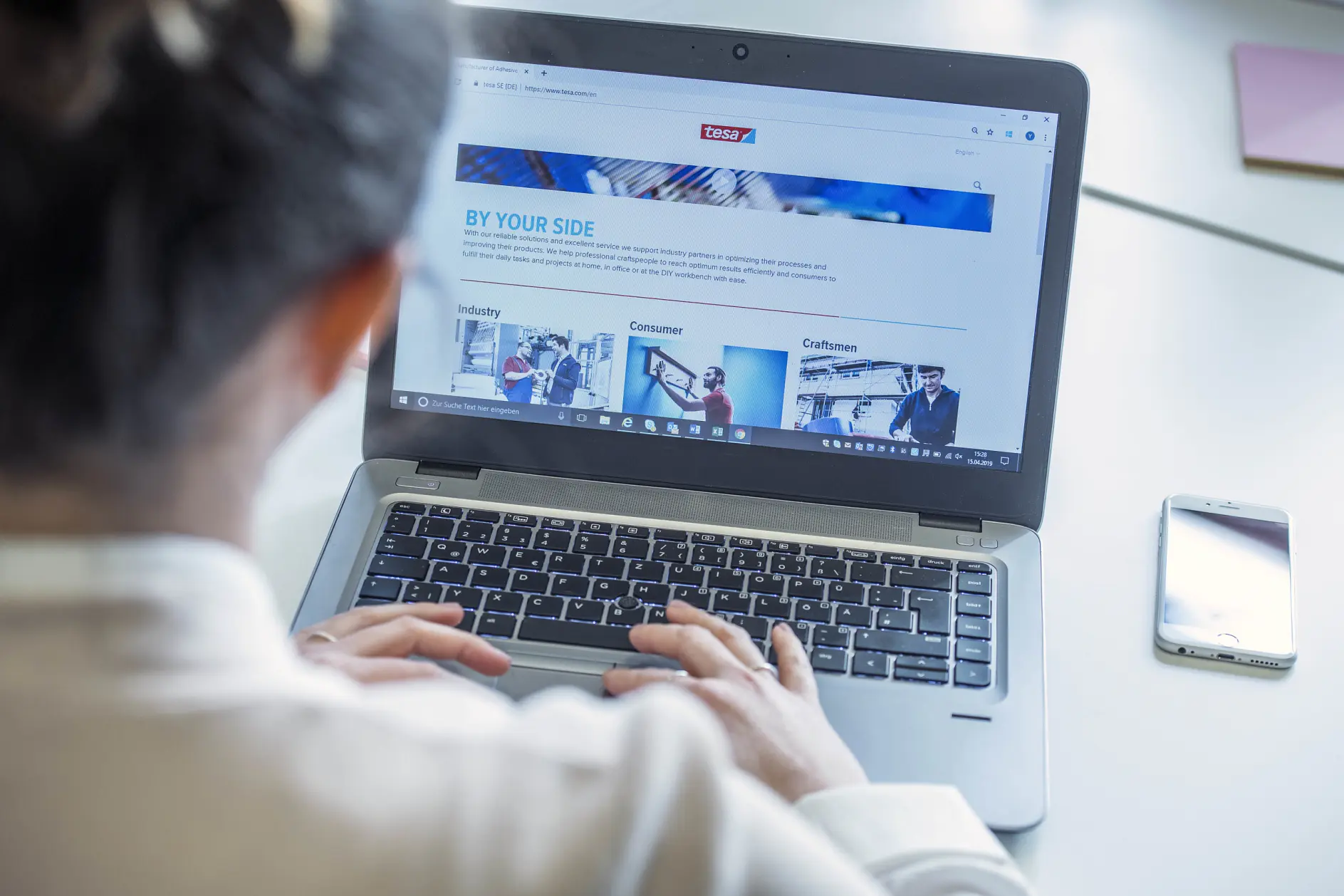 A person uses a laptop displaying a webpage titled BY YOUR SIDE with images and text. The laptop is placed on a white surface next to a smartphone, accompanied by tesa tape. The person is wearing a white shirt and has short hair.