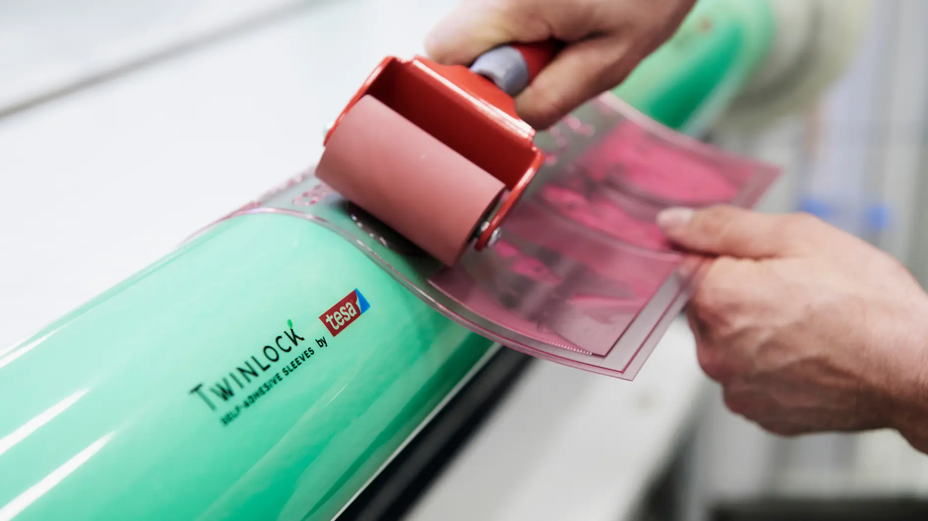 Close-up of hands using a small roller to apply a pink sheet onto a large green cylinder labeled Twinlock. The cylinder also displays the phrase Self Adhesive Sleeves, with tesa tape being used in the process.