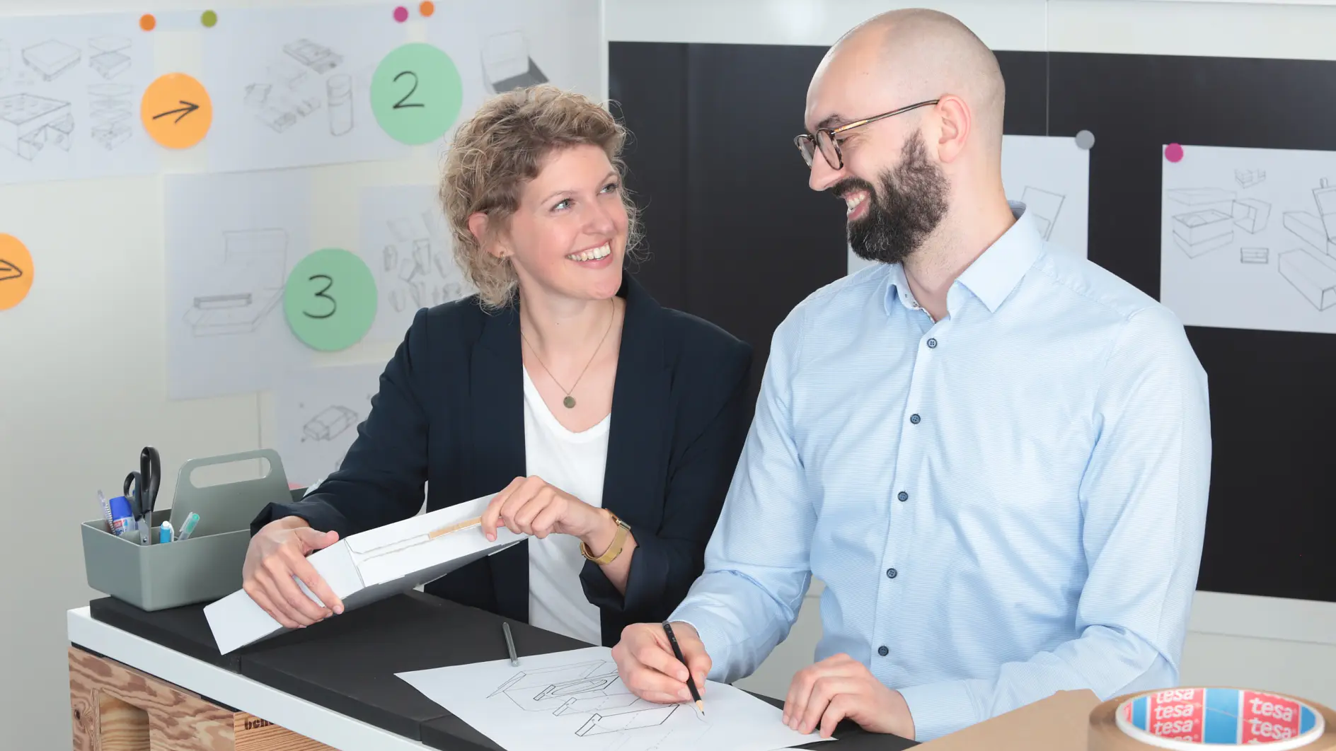 Two people, a woman and a man, are working together at a desk with design papers, models, and sketches. They are smiling and surrounded by office supplies and design drawings on the wall. A tray of stationery is on the desk, featuring tesa tape prominently among other items. (This text has been generated by AI)